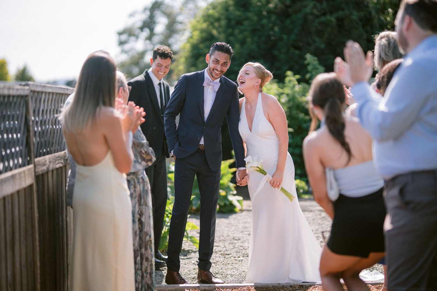 Smiling newlyweds walk through cheering guests after outdoor wedding ceremony on sunny day.