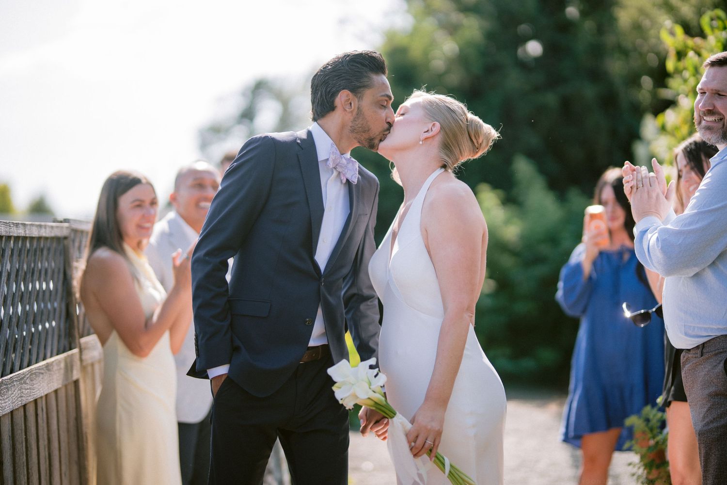 Newlyweds sharing first kiss surrounded by cheering guests during sunny outdoor wedding celebration.