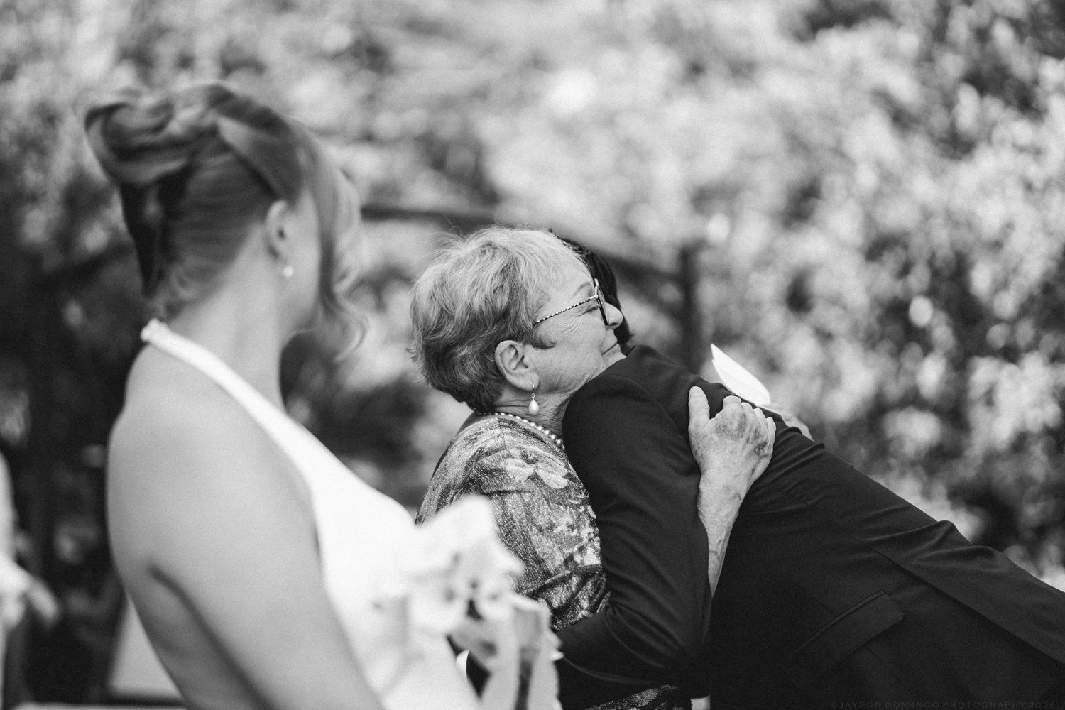 Black and white photo of an emotional family member embracing the groom during a wedding ceremony.