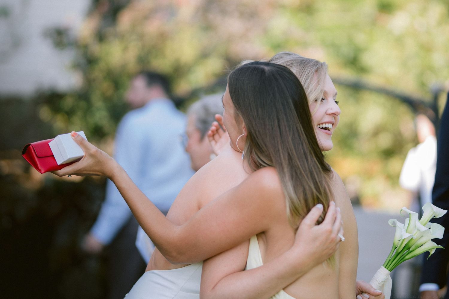 Two women sharing a warm hug while one holds a small red gift box at a wedding reception.