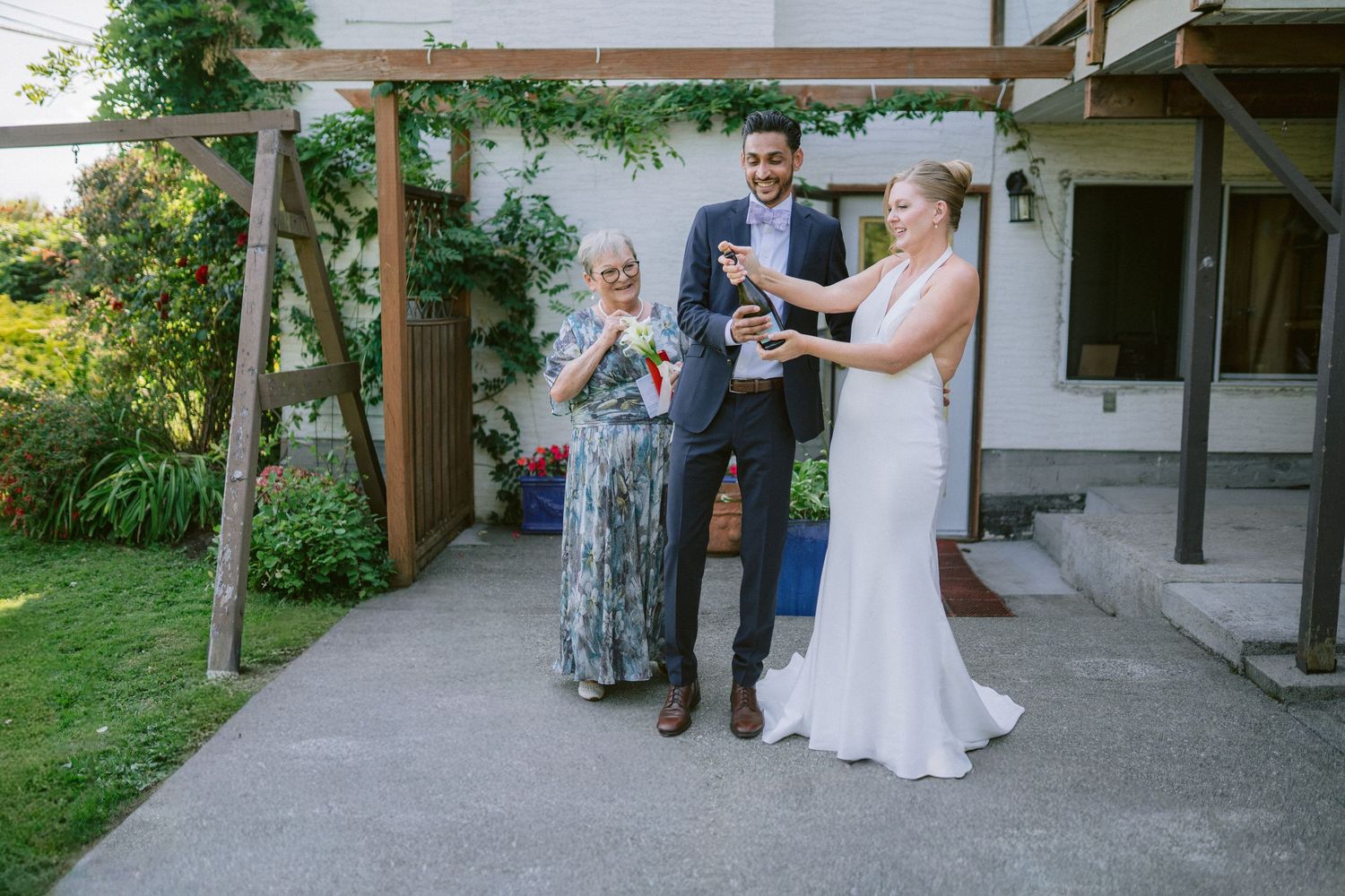 Newlywed couple celebrates with champagne alongside smiling older woman in garden wedding setting.