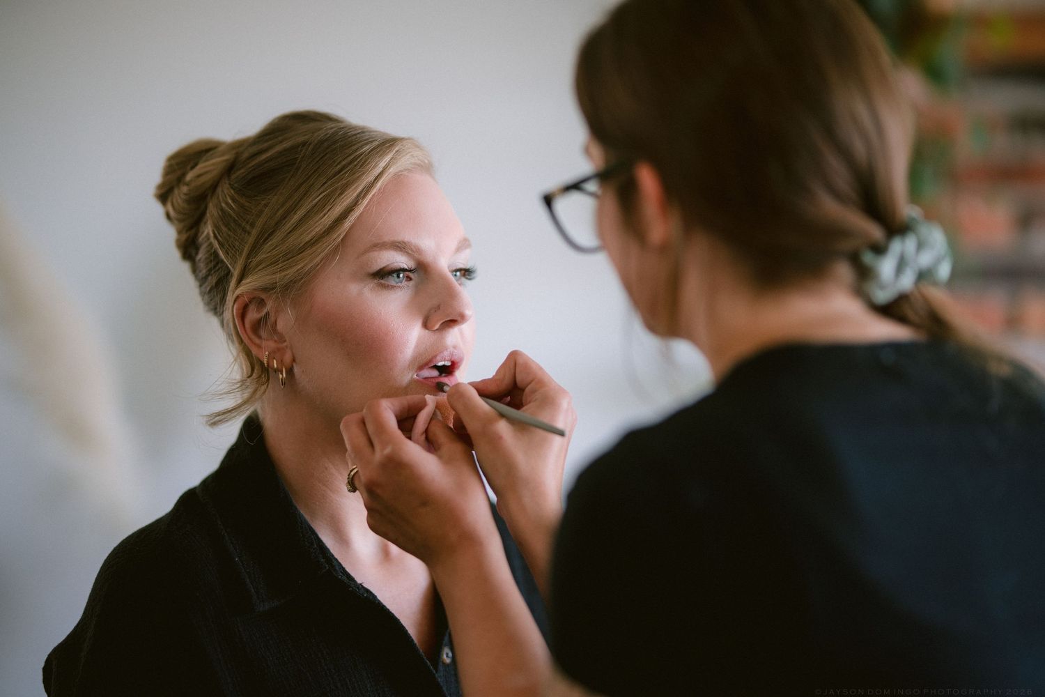 Makeup artist applying lipstick to blonde bride during bridal beauty preparation session.