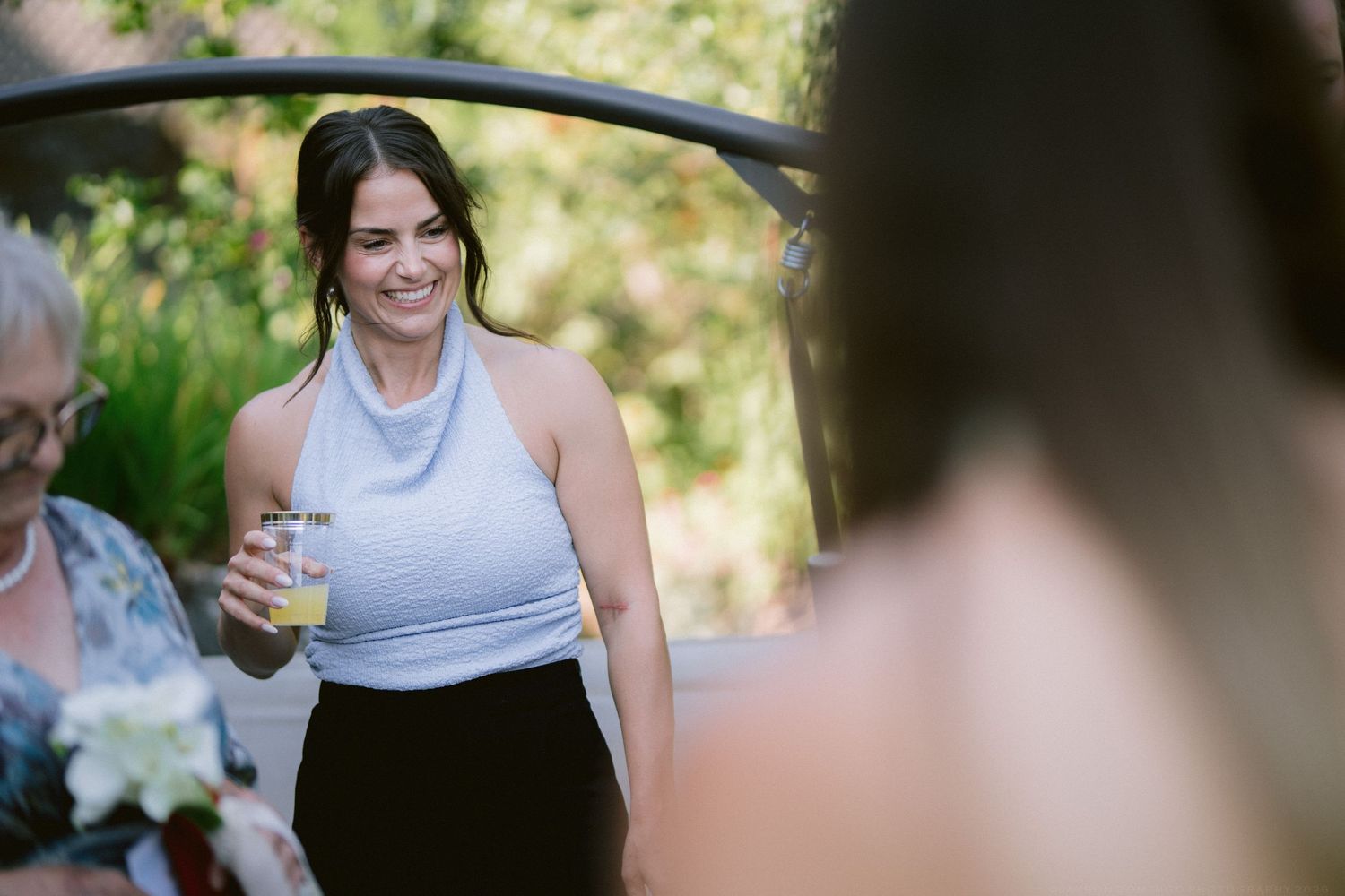 Smiling dark-haired woman in light blue top holding a drink at an outdoor wedding celebration.