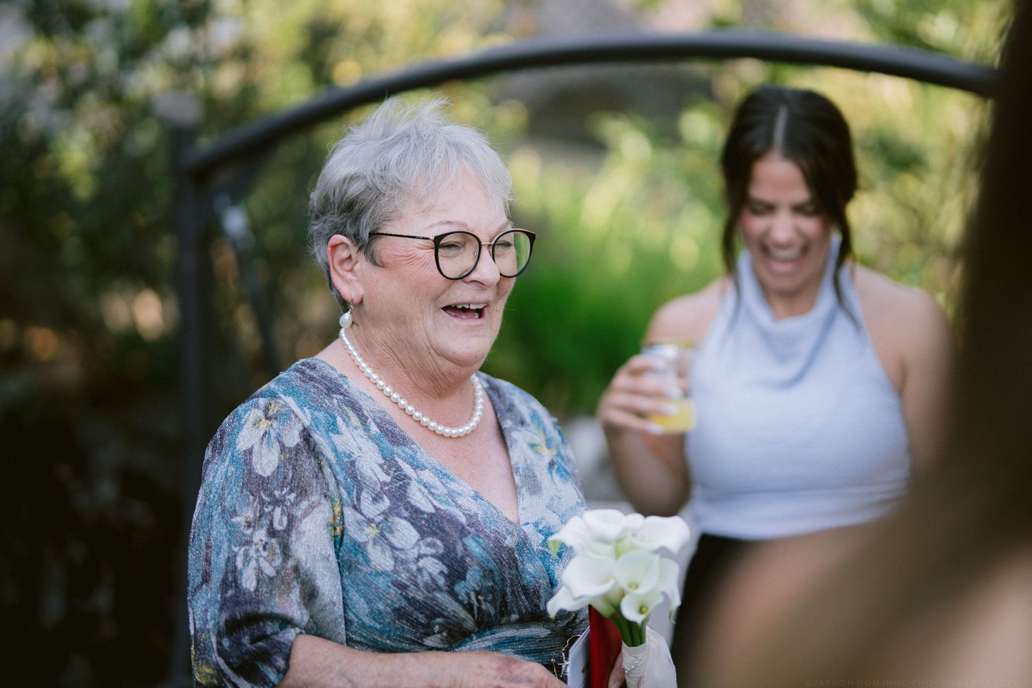 Joyful older woman with glasses and pearl necklace laughing while holding white flowers at wedding.