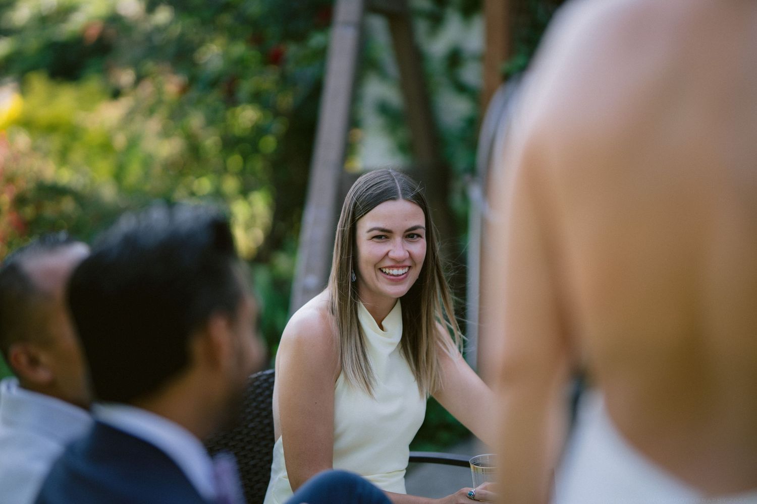 Smiling woman in yellow dress enjoys outdoor wedding reception surrounded by guests and greenery.