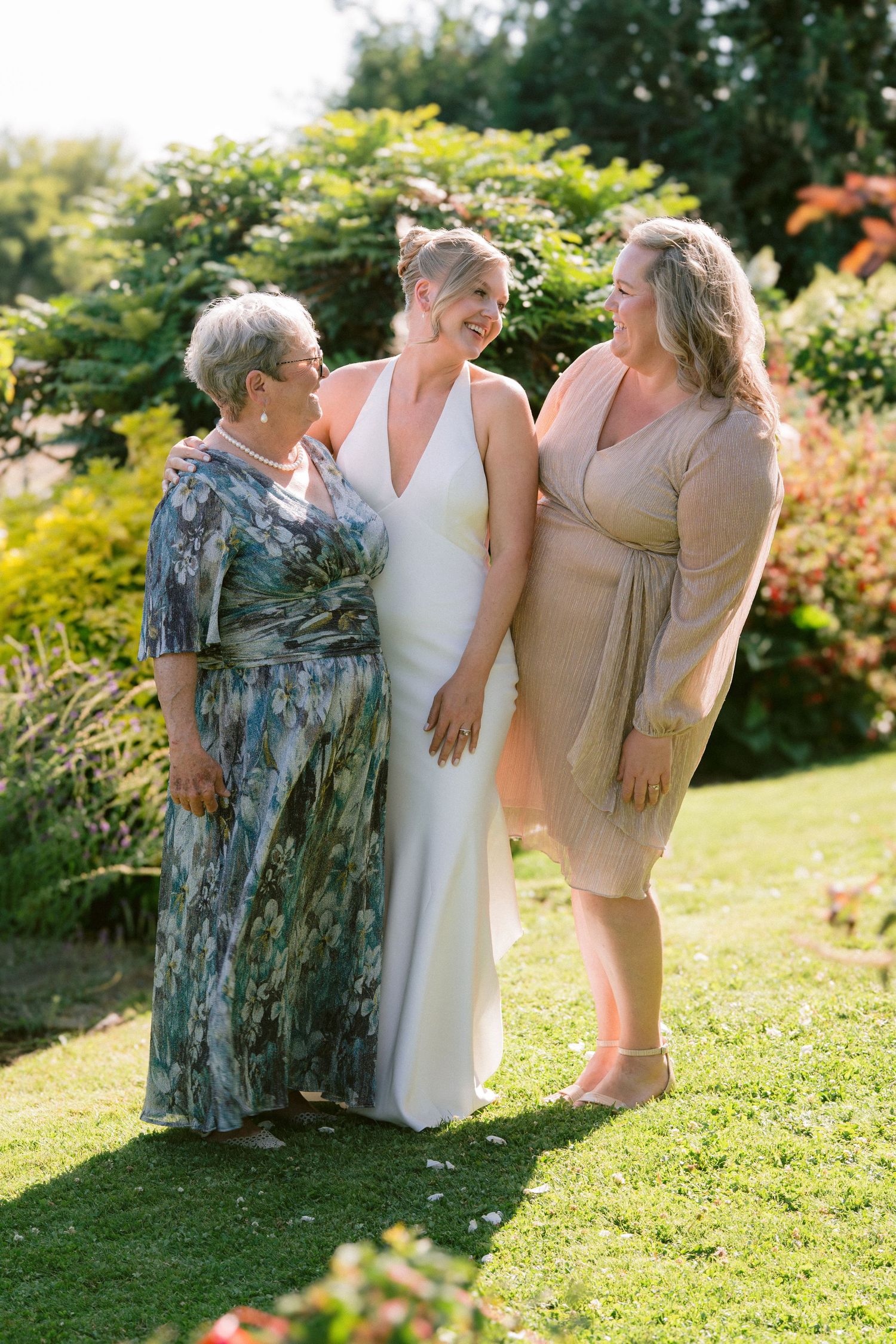 Bride in white halter gown laughing with two female family members in sunny garden setting.