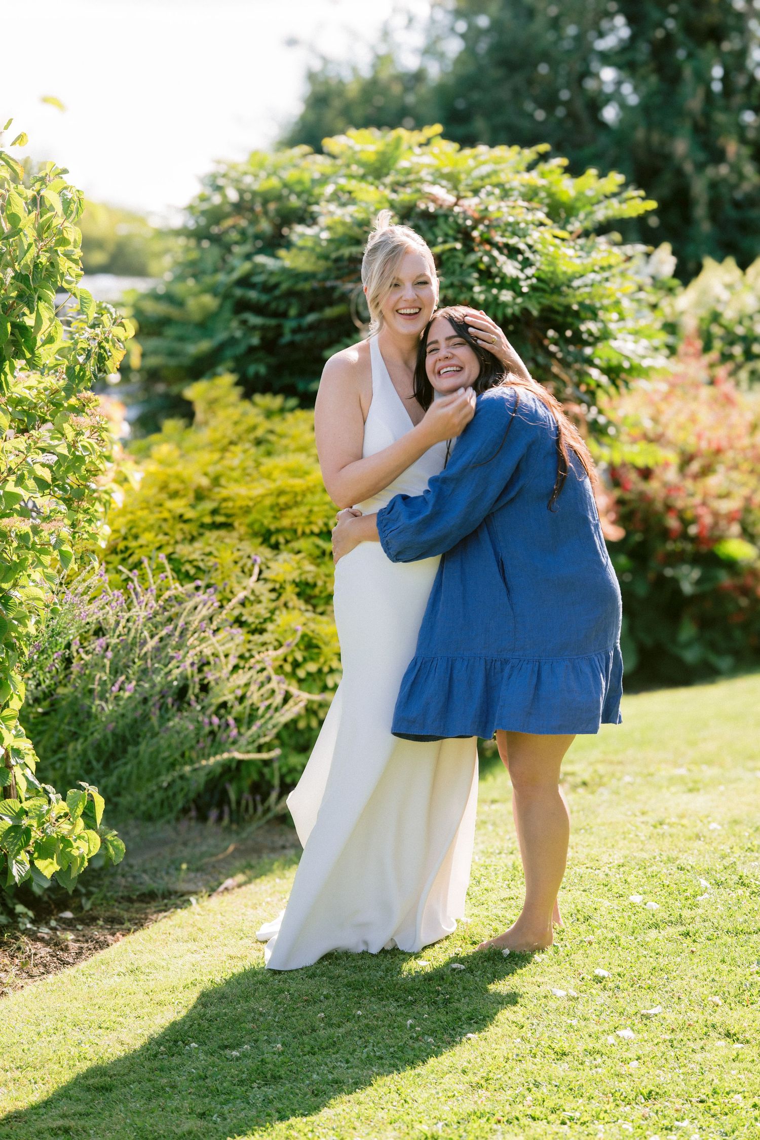 Bride in white halter gown joyfully embracing a guest in blue dress in sunny garden.