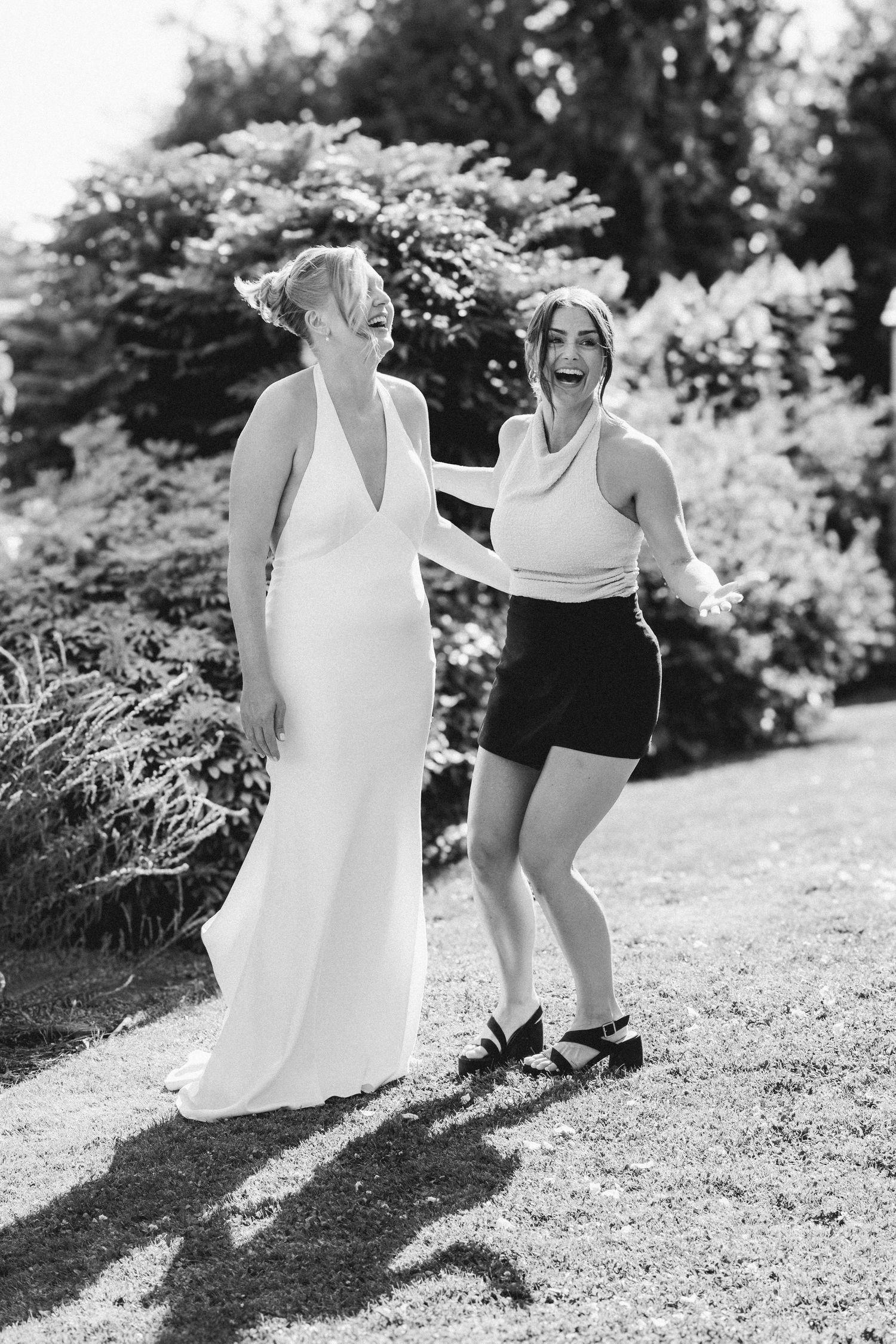 Bride in white gown laughing with friend in black and white outdoor wedding photo.