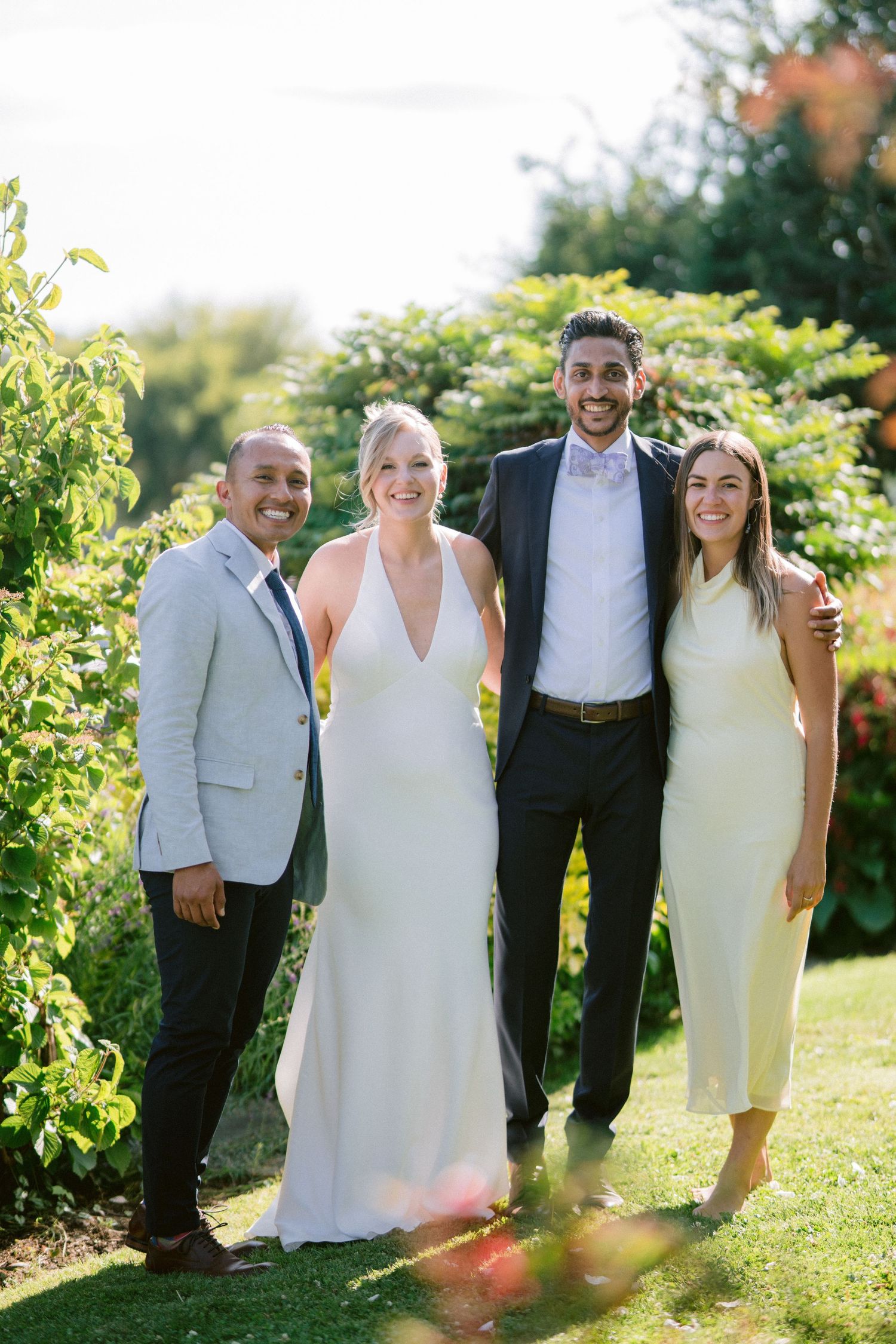 Bride and groom pose with two guests in garden setting at sunny outdoor wedding reception.