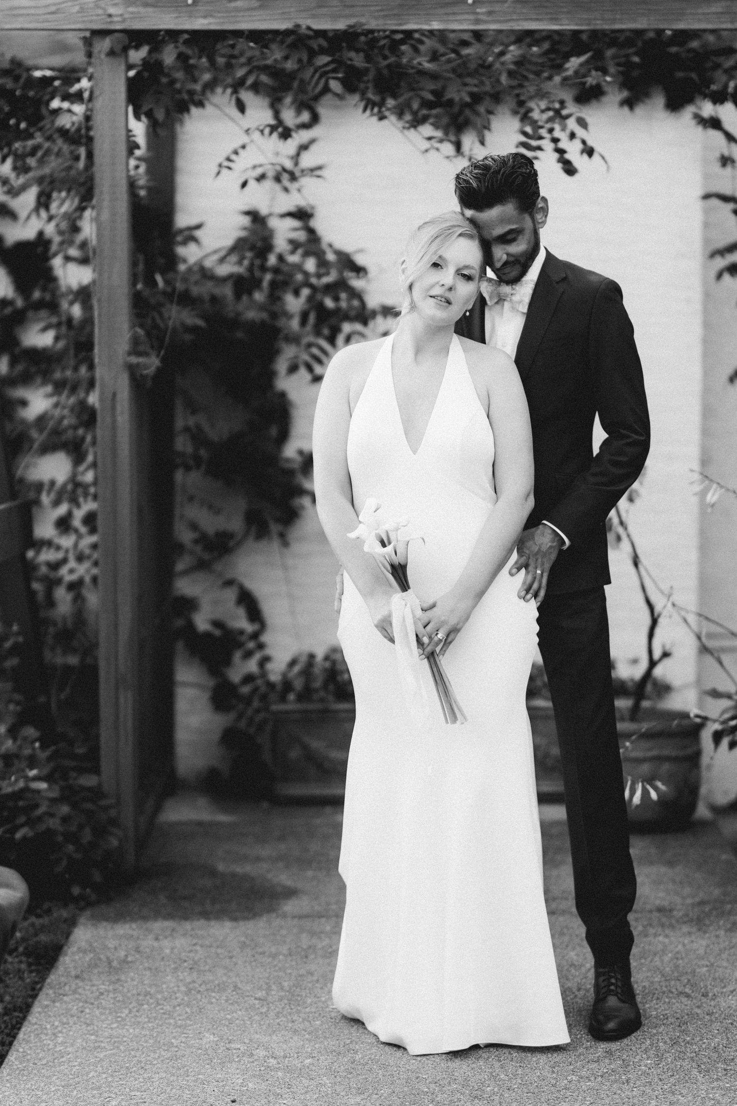 Black and white portrait of newlywed couple embracing under vine-covered pergola after wedding ceremony.