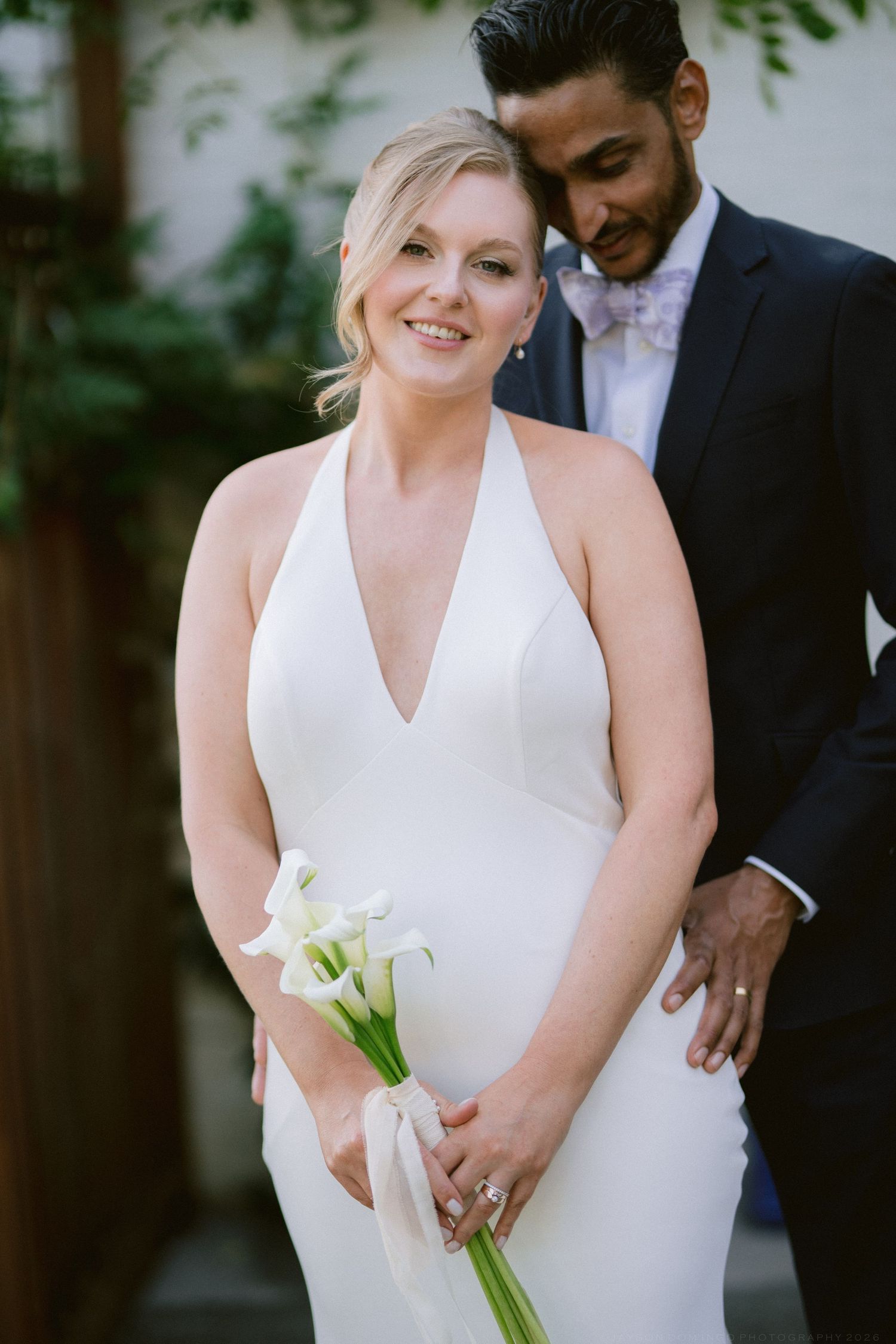 Smiling bride in halter wedding gown holding calla lily bouquet with groom in navy suit behind her.