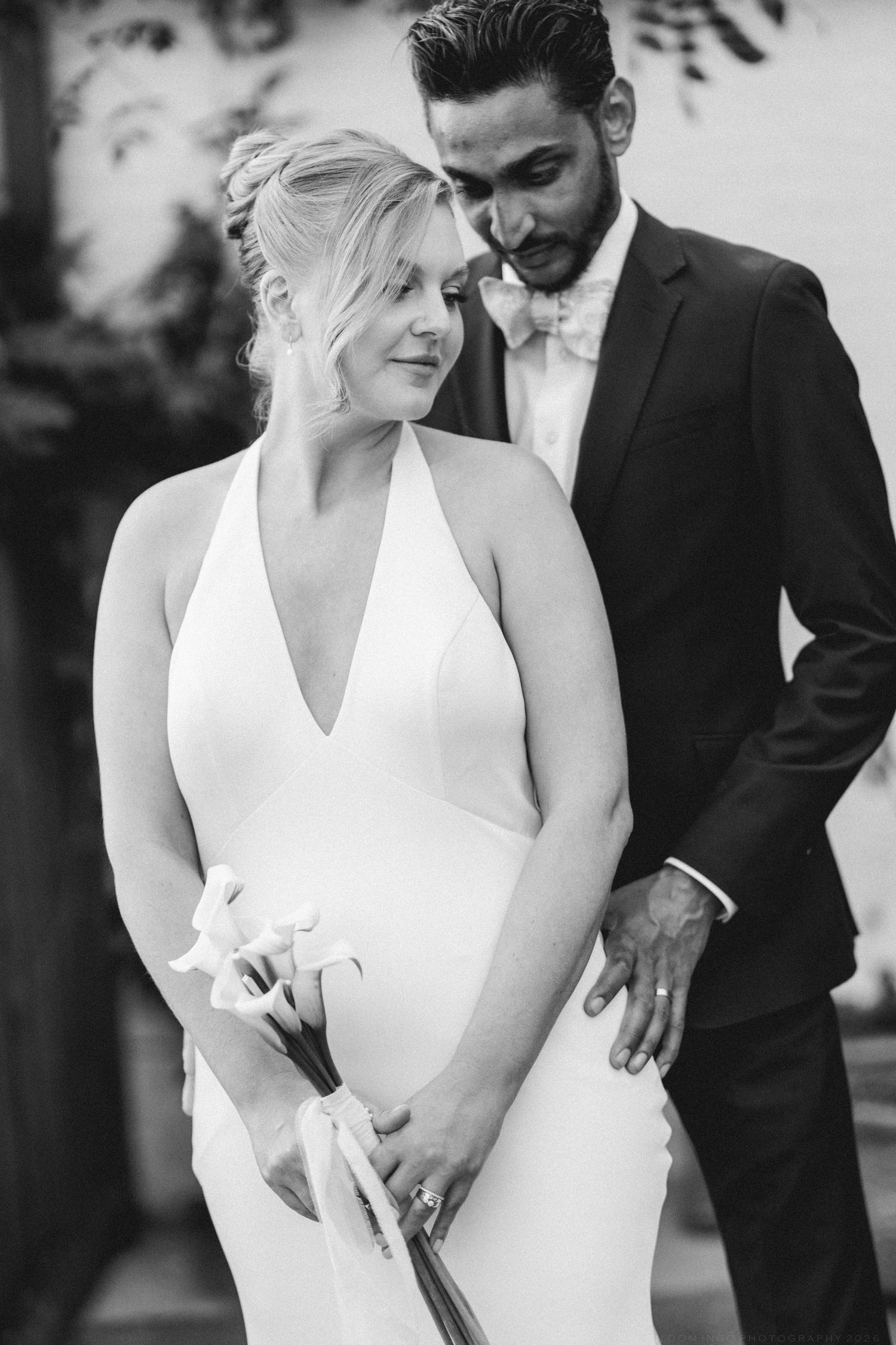 Bride in elegant halter gown holding flowers with groom in suit, black and white wedding portrait.