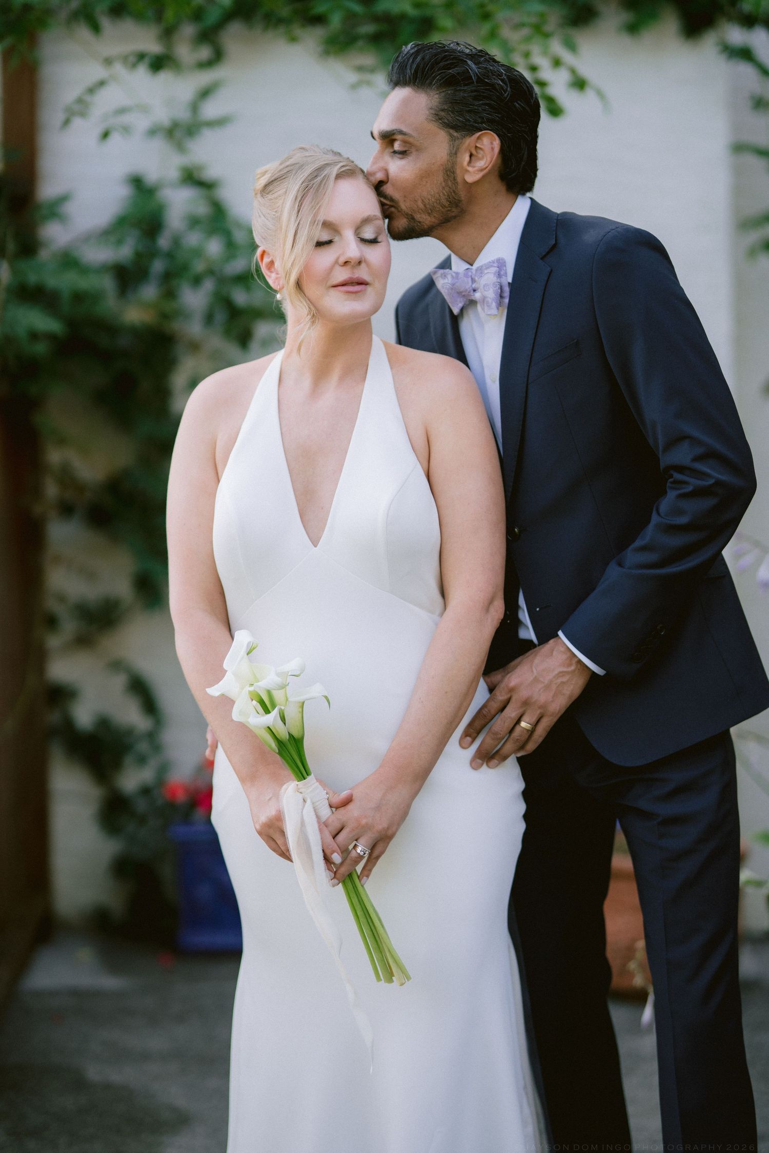 Groom in navy suit kissing bride on temple while she holds white calla lily wedding bouquet.