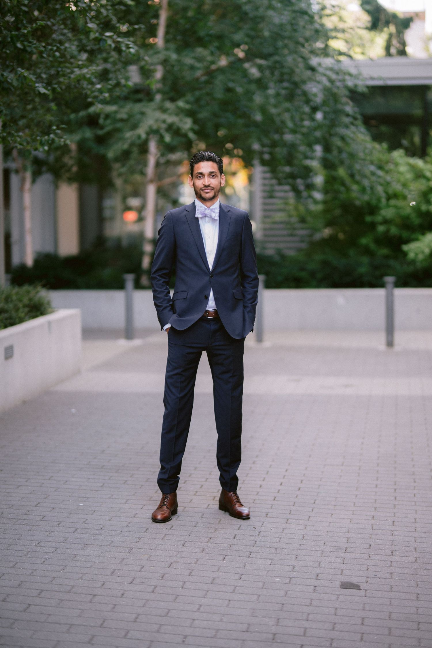 A young man in a navy blue suit and brown dress shoes stands confidently on a brick plaza surrounded by greenery.