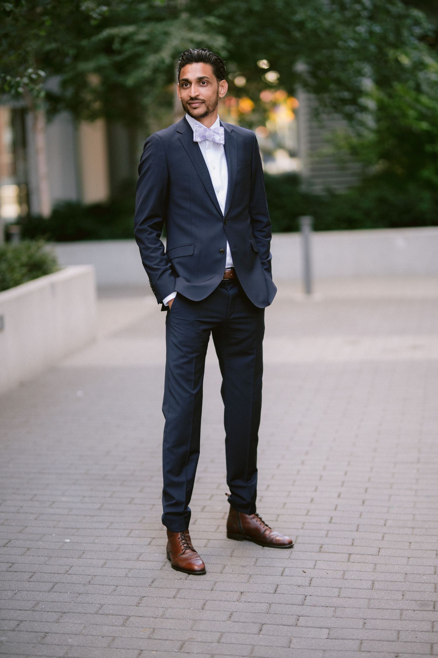 Dapper groom in navy suit with pink bow tie and brown oxford shoes posing outdoors on urban brick pathway.