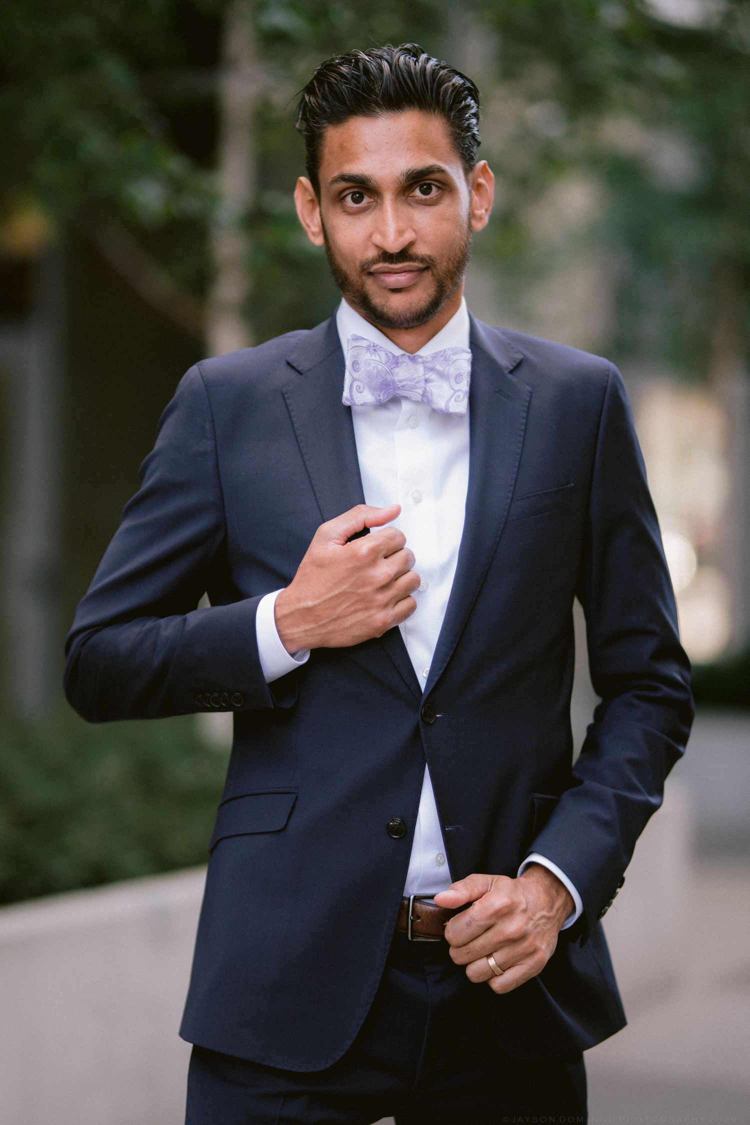 A well-dressed man in a navy suit and lavender bow tie poses confidently outdoors with greenery in the background.