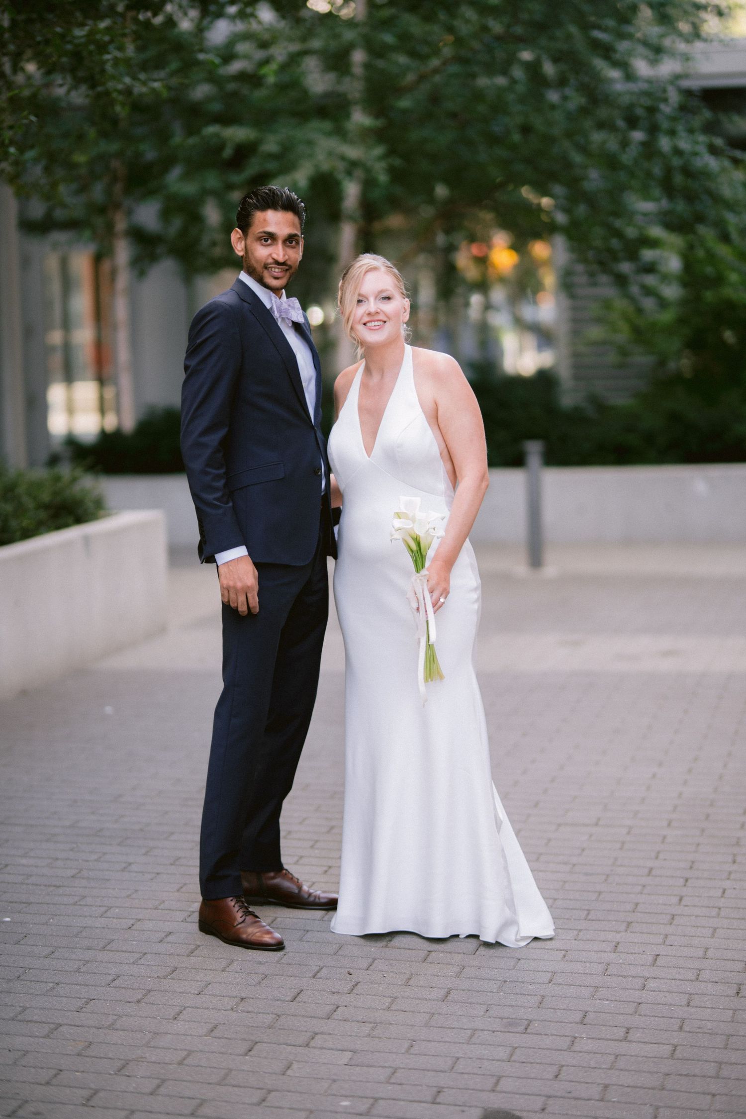 Bride and groom posing together outdoors on brick pathway, bride in white halter gown holding calla lilies, groom in navy suit.