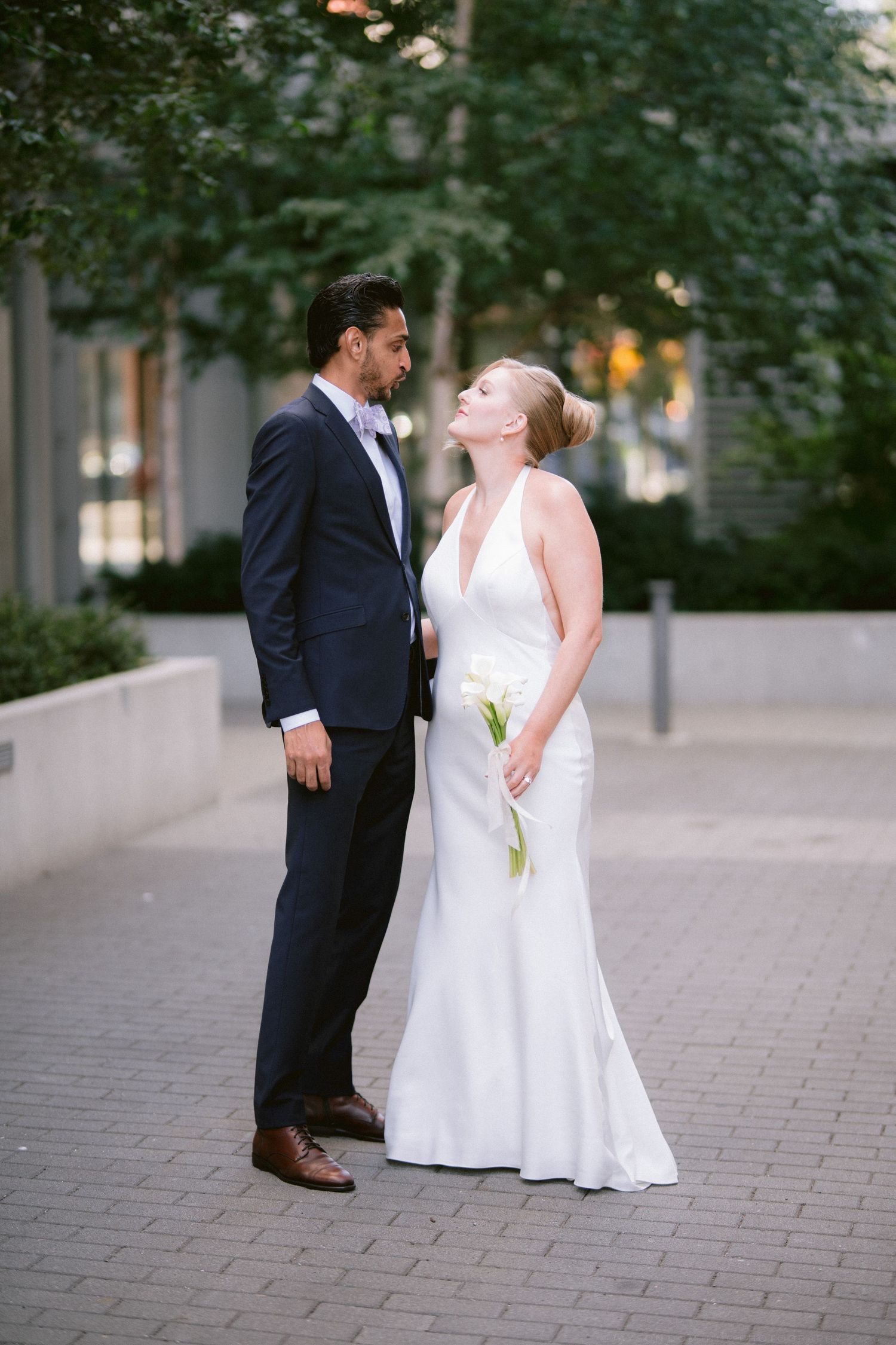 Bride and groom share a romantic moment outdoors on a cobblestone path, she in a white halter gown holding calla lilies.