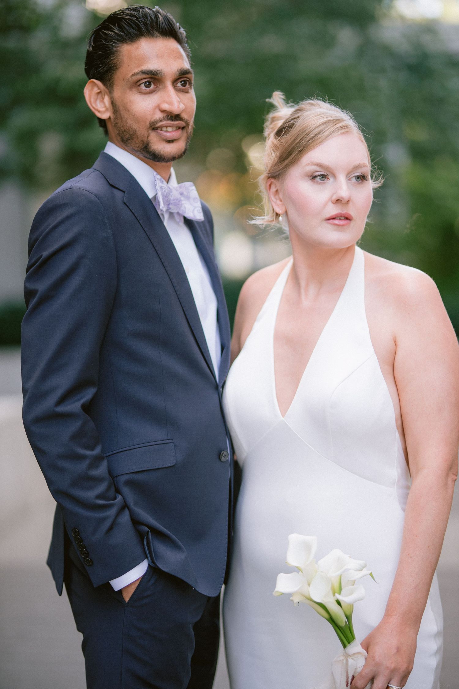 Interracial wedding couple posing outdoors, groom in navy suit with bow tie, bride in white halter gown holding calla lilies.