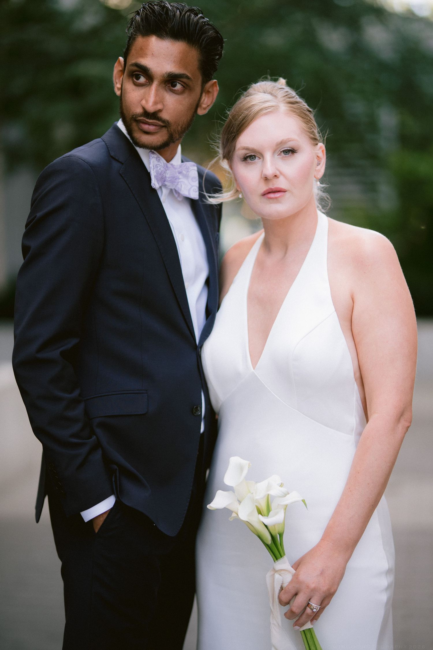 Interracial couple in wedding attire, groom in navy suit with lavender bow tie, bride in white halter gown holding calla lilies.