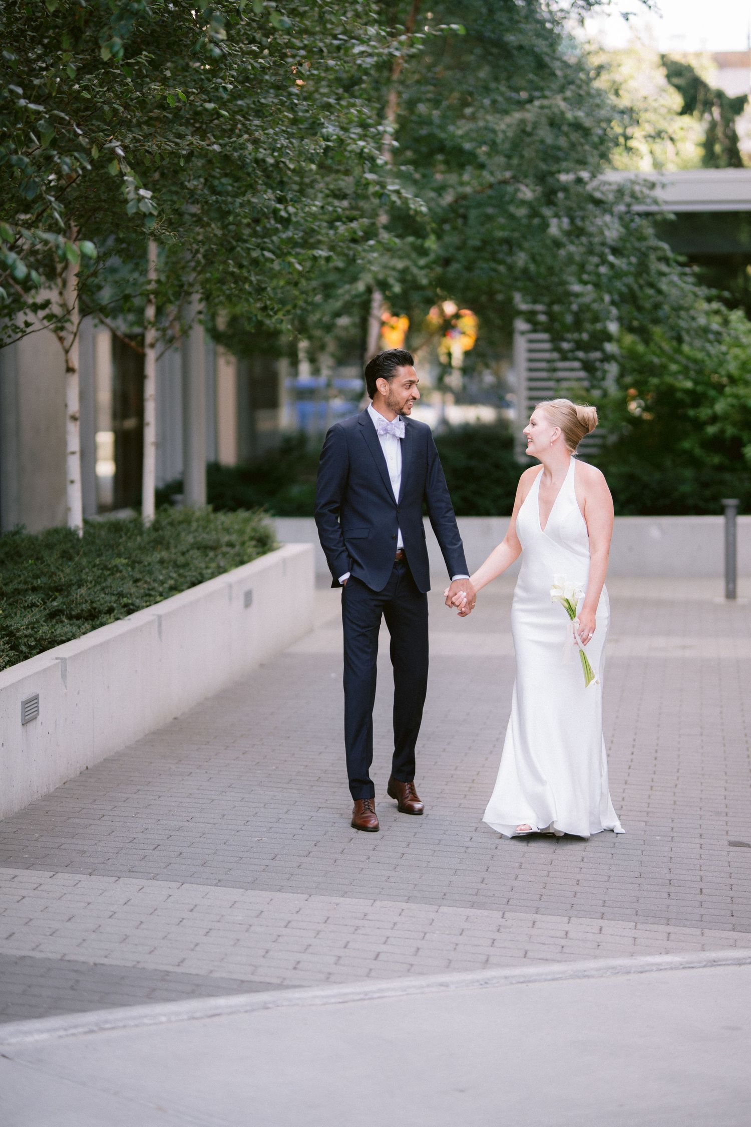 Bride and groom walking hand in hand on urban brick pathway, smiling at each other, bride in white gown, groom in navy suit.
