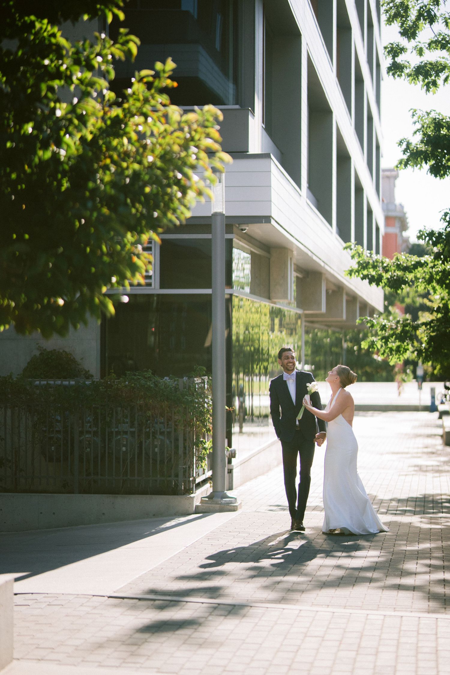 A bride and groom walk hand in hand along a sunny urban sidewalk beside a modern glass building.