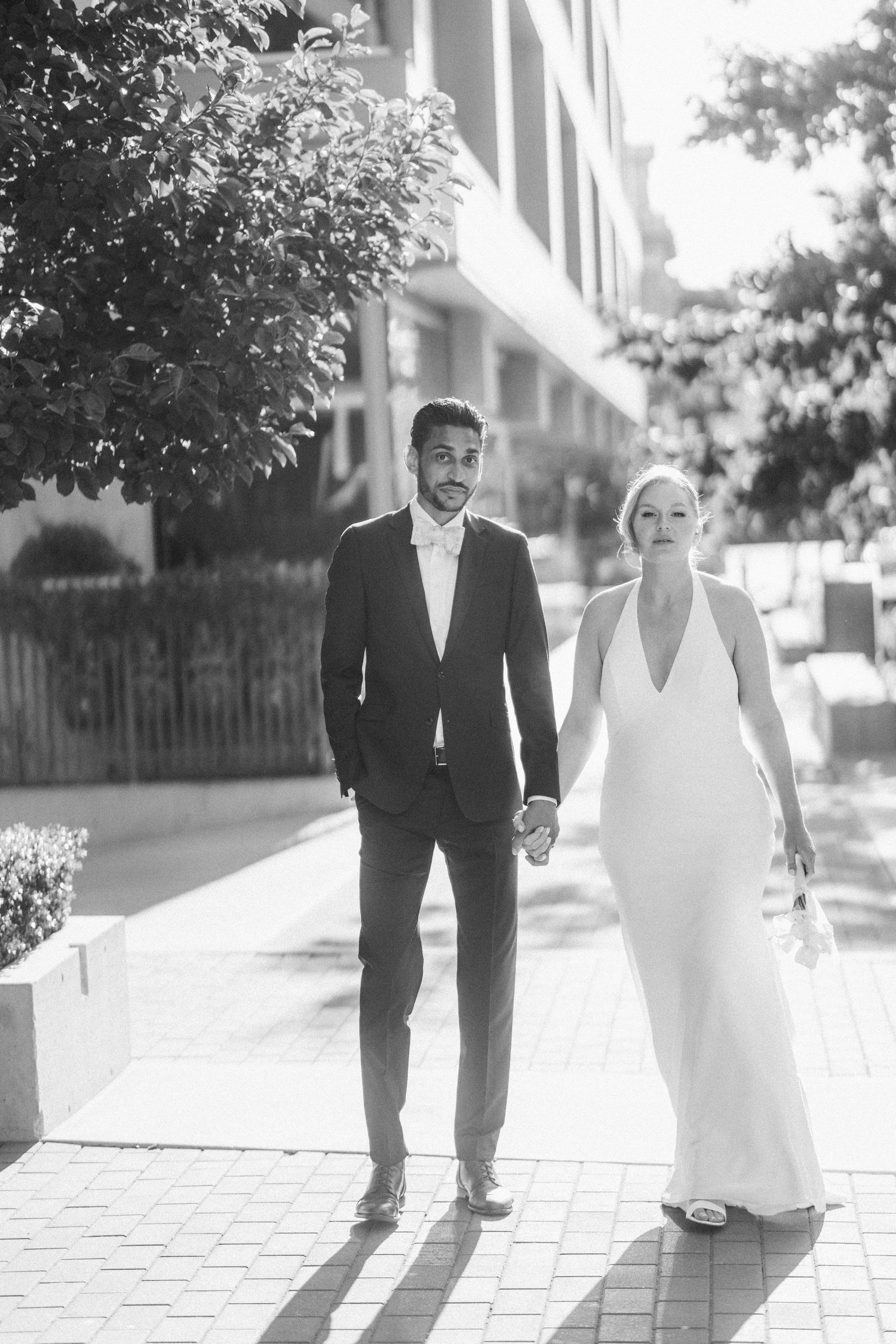 Black and white photo of a bride and groom holding hands walking on a sunny urban sidewalk in elegant wedding attire.