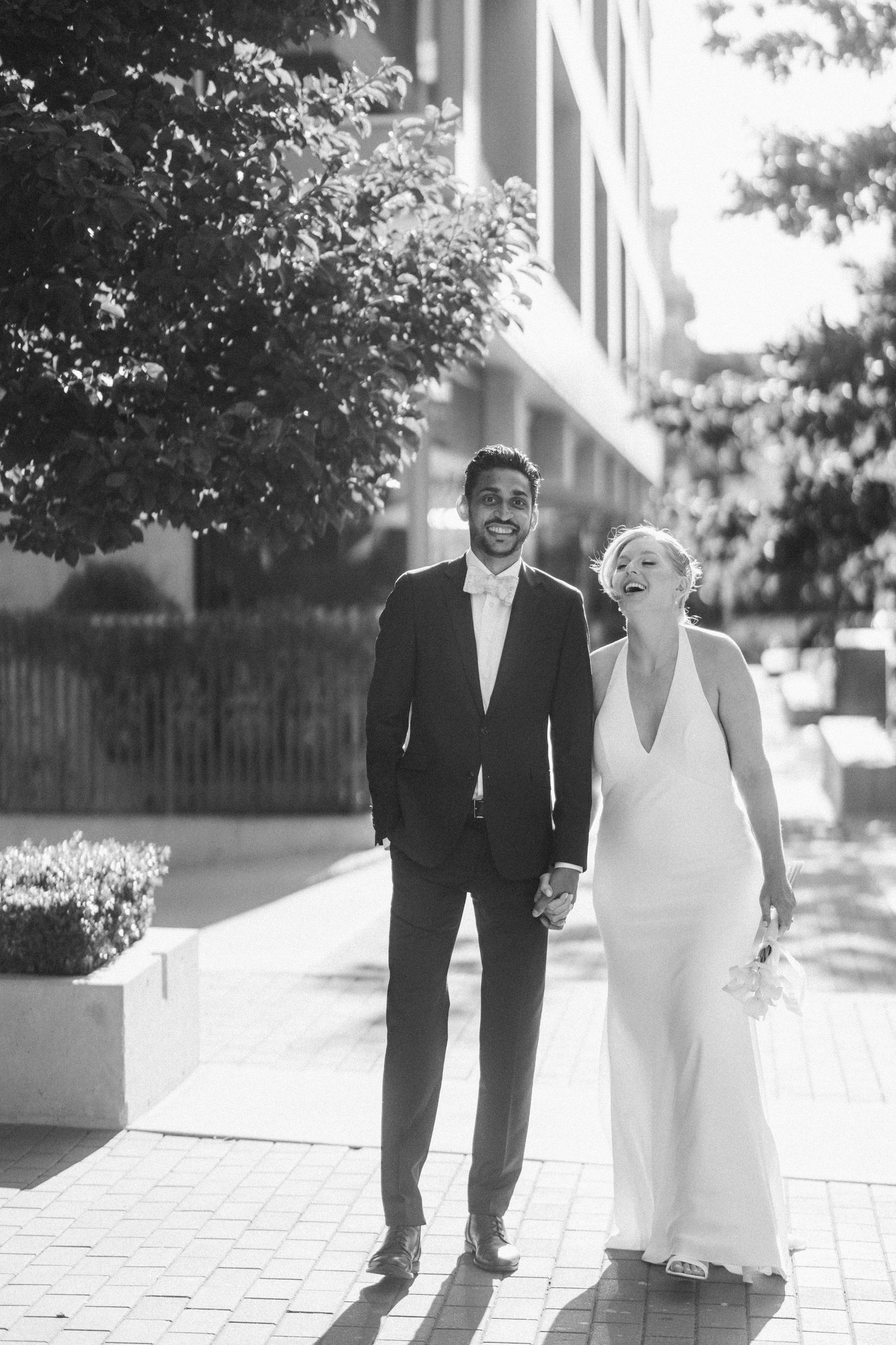 Black and white photo of a laughing bride and groom walking hand in hand on a sunny urban sidewalk.