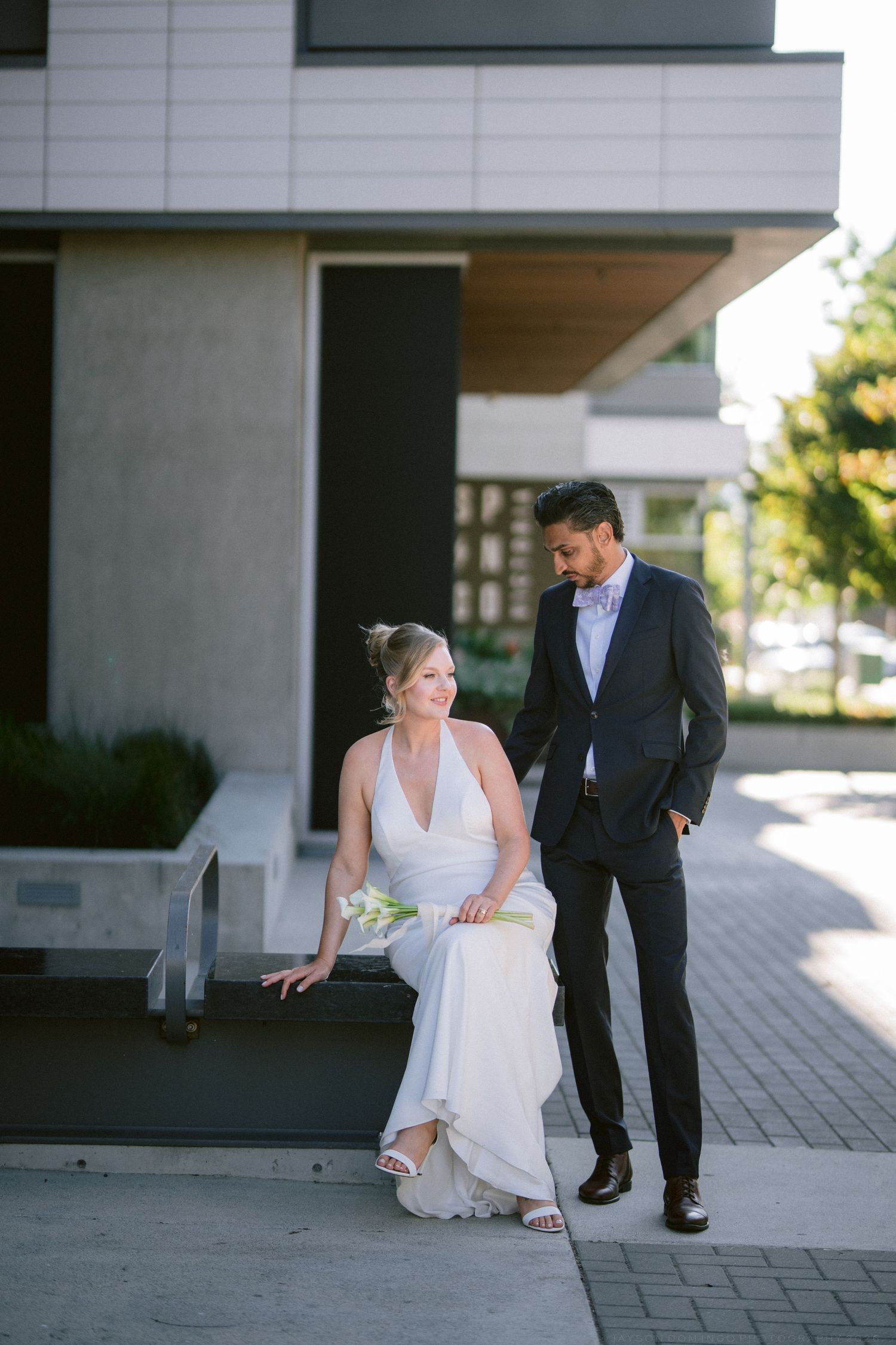 Bride in white halter gown sits outdoors with groom in navy suit at modern building during wedding photos.