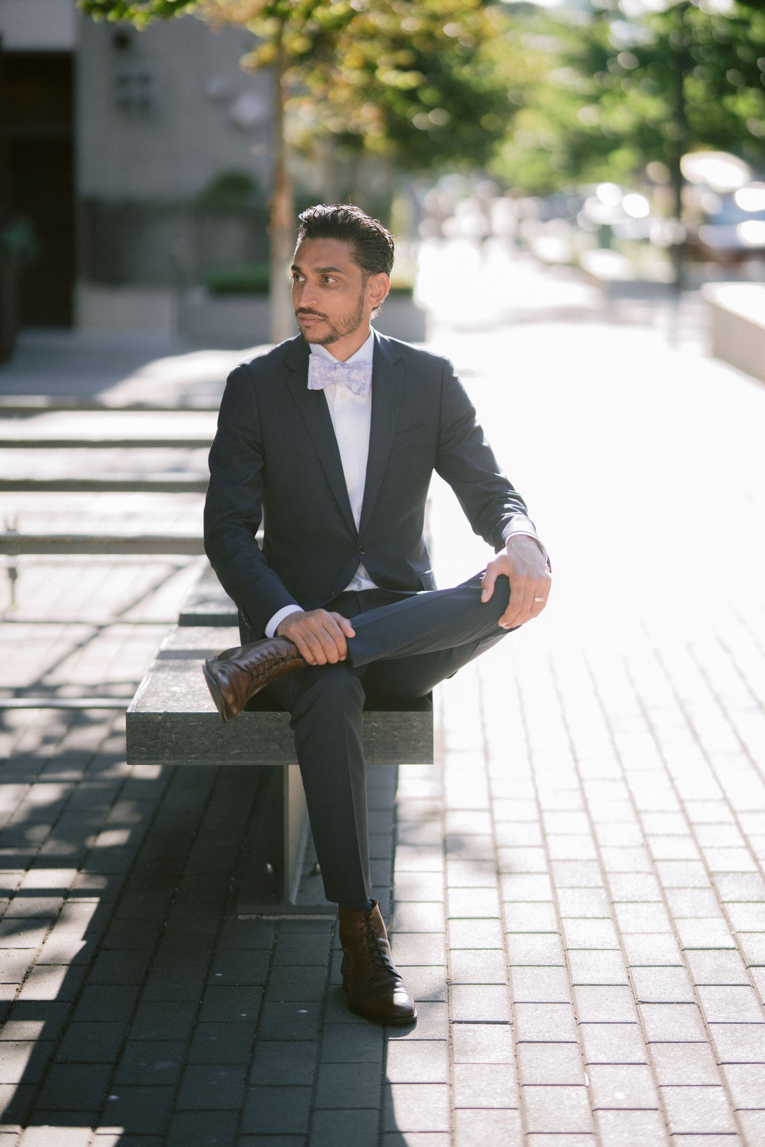 A well-dressed man in a dark suit and bow tie sits on a concrete bench outdoors in an urban setting.