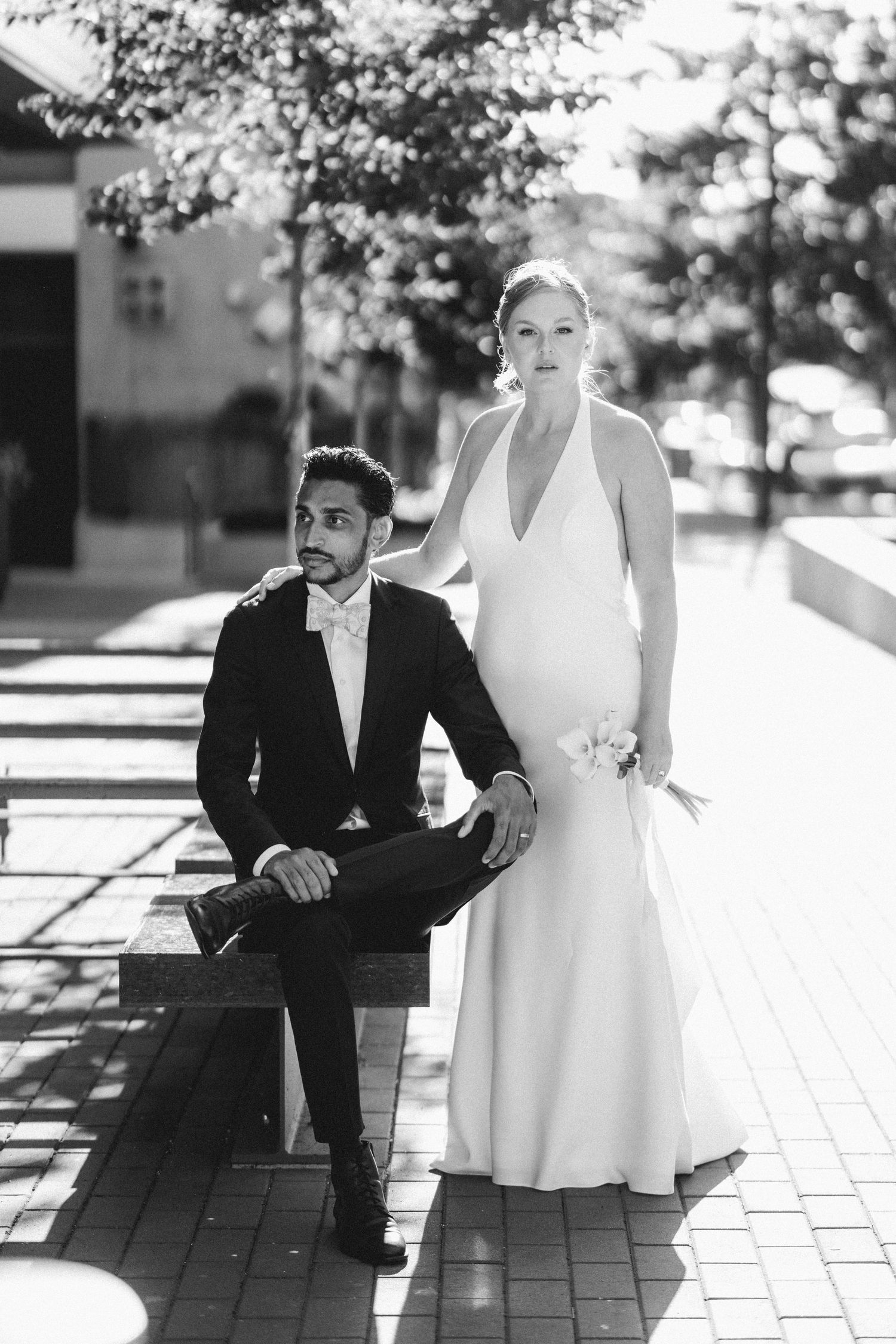 Black and white photo of wedding couple, groom seated on bench in suit, bride standing beside him in elegant halter gown.