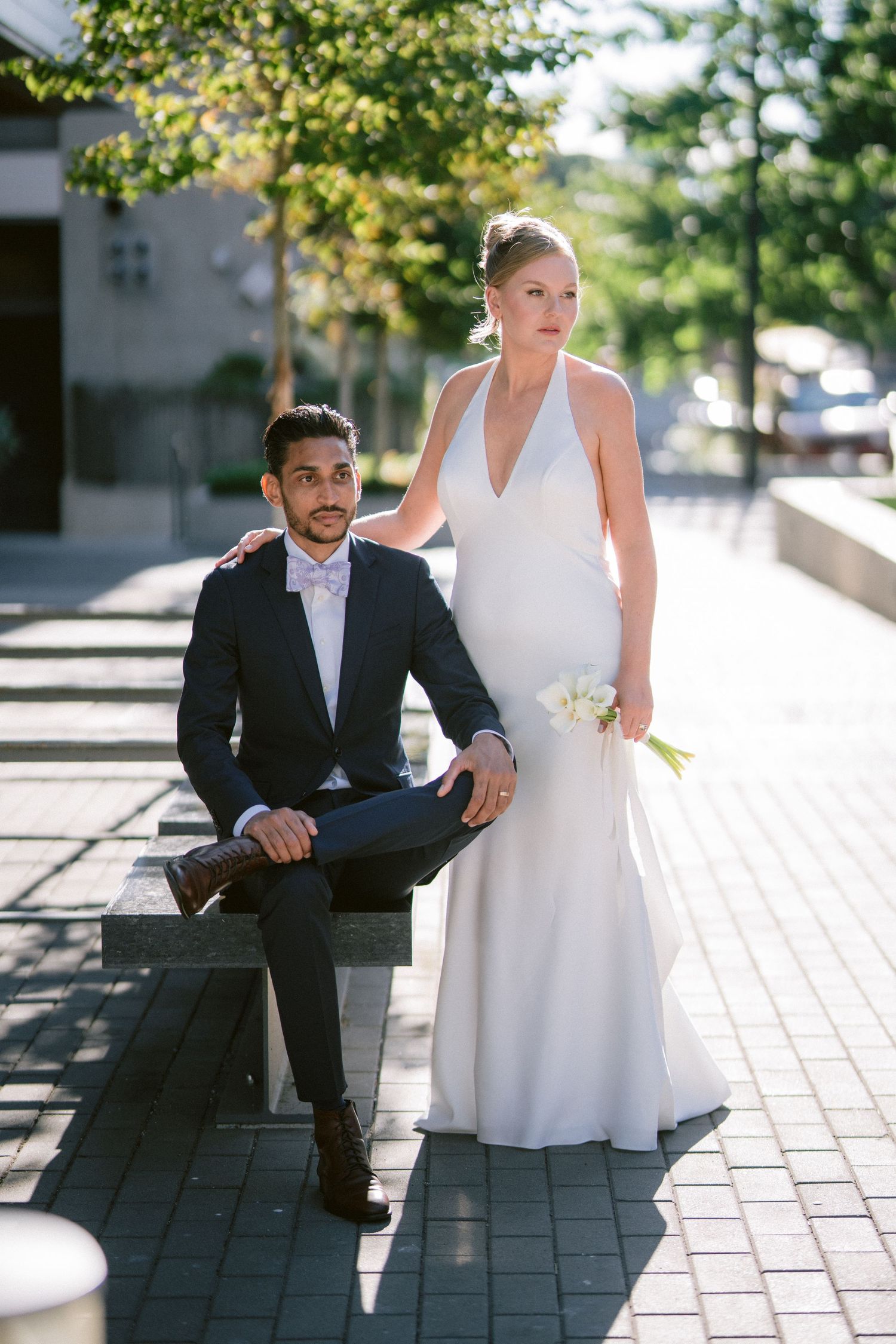 Bride in white halter gown and groom in navy suit pose together on urban bench during wedding photo session.