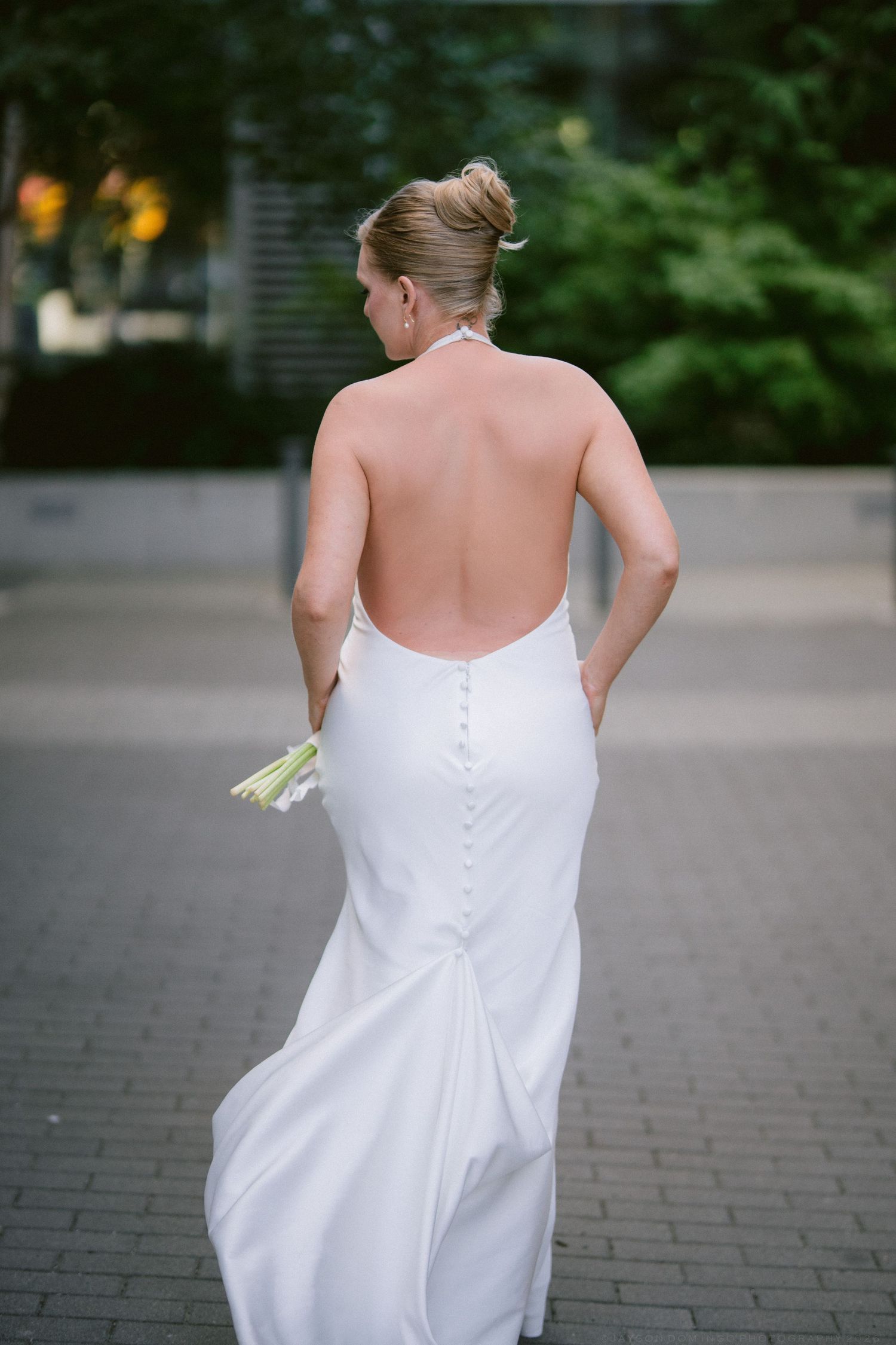 Bride in elegant backless white halter wedding gown holding bouquet of white flowers on outdoor paved walkway.