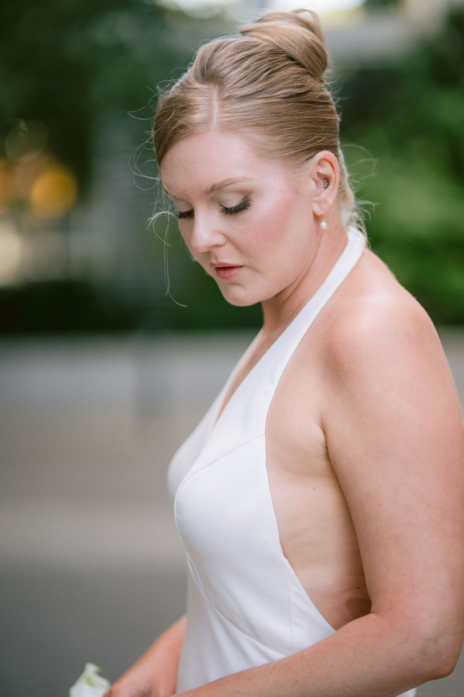 Elegant bride with updo hairstyle in white halter wedding gown looking down gracefully outdoors.