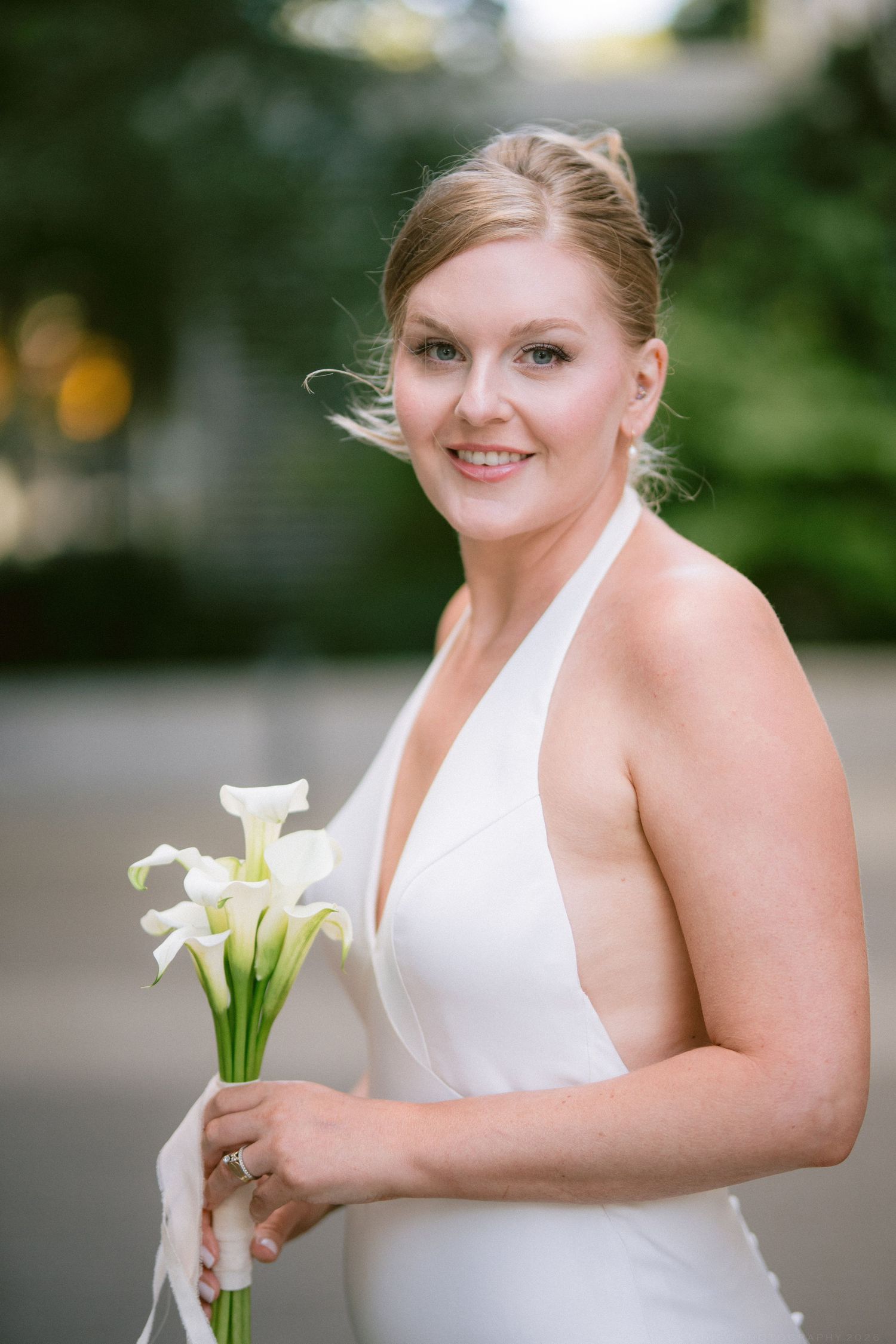 Smiling bride in a white halter wedding gown holds a bouquet of white calla lilies outdoors.