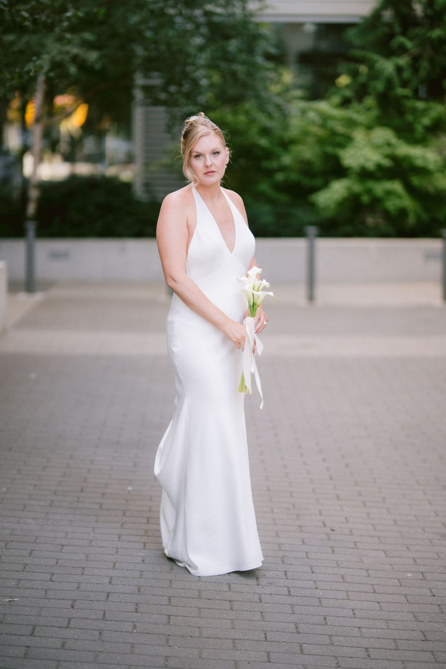 Elegant bride in white halter neck wedding gown holding calla lily bouquet on cobblestone path.