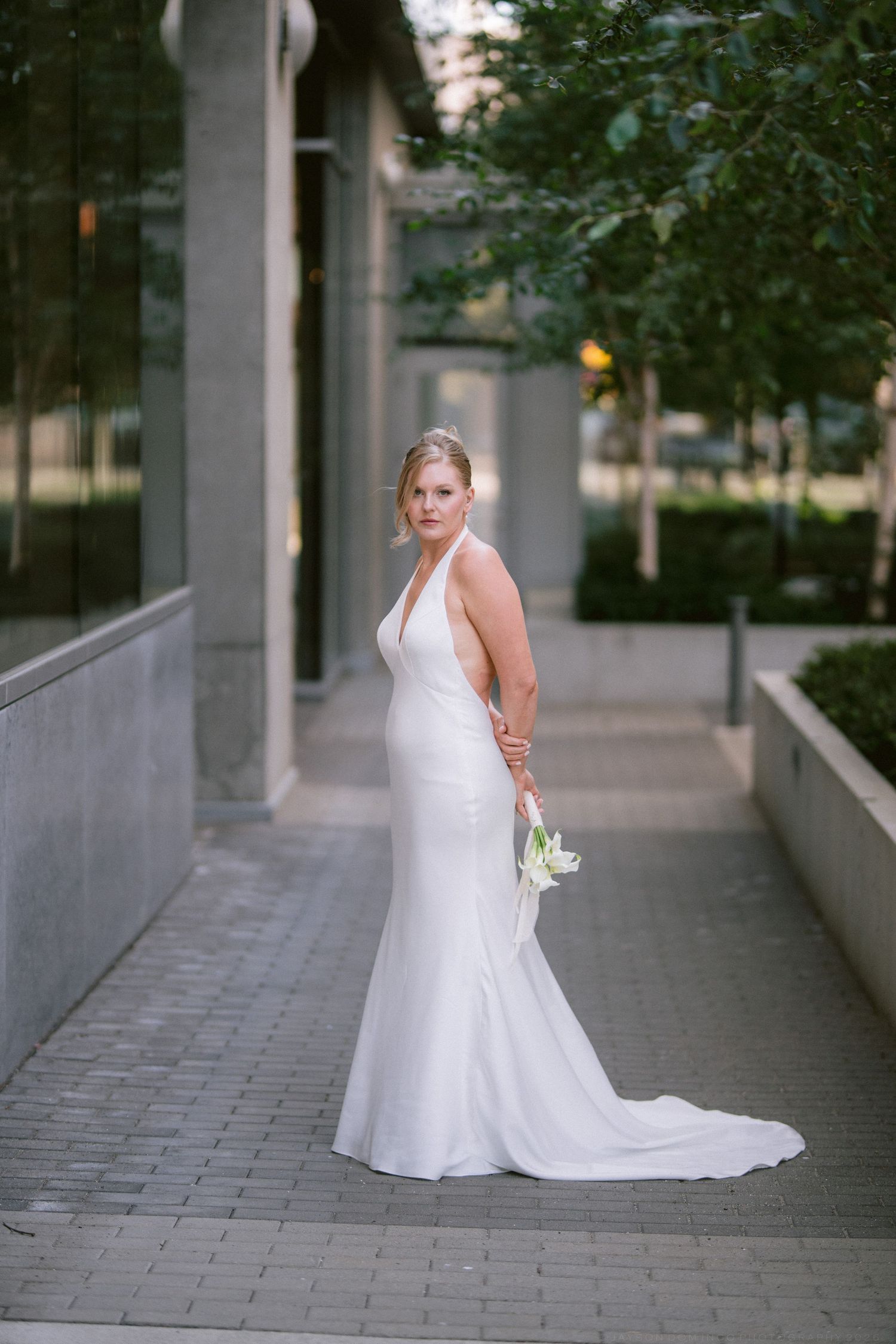 Elegant bride in a fitted white halter mermaid gown poses on a cobblestone walkway with calla lilies.