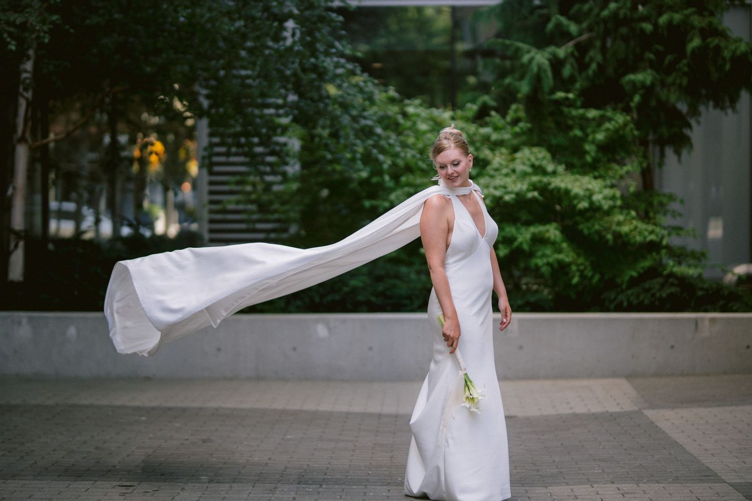 Bride in white halter gown with flowing cape veil poses outdoors with lush green trees in background.