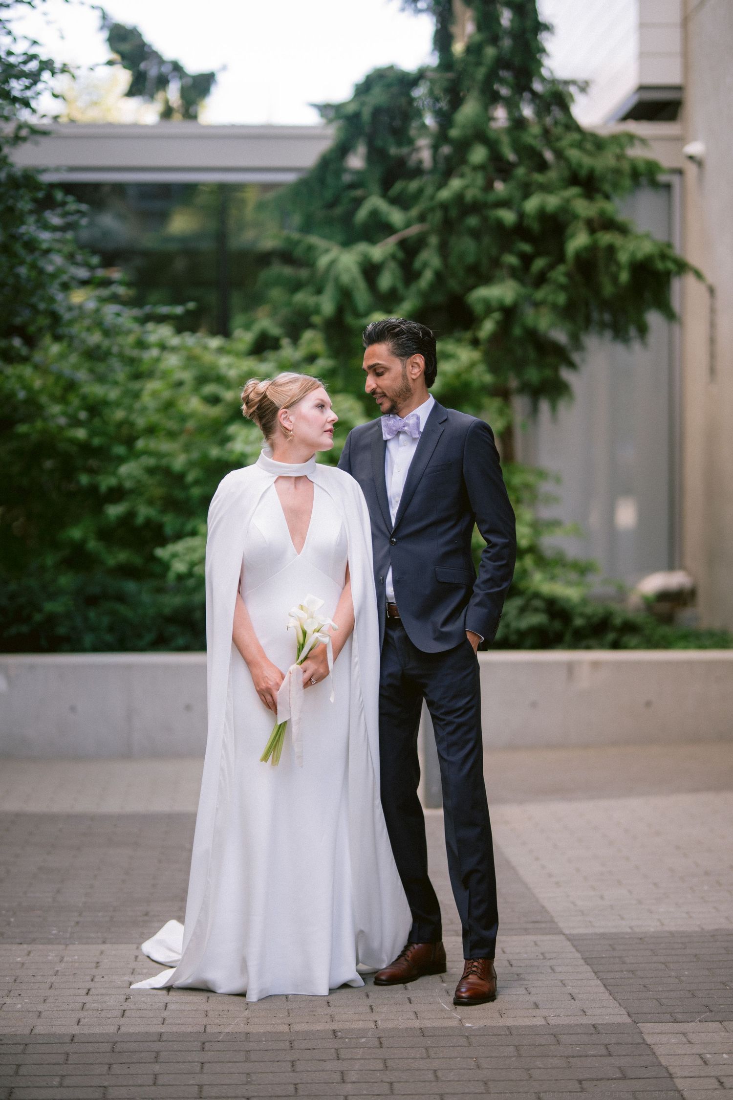 Bride in a white cape gown and groom in navy suit share a loving glance outdoors with lush greenery.