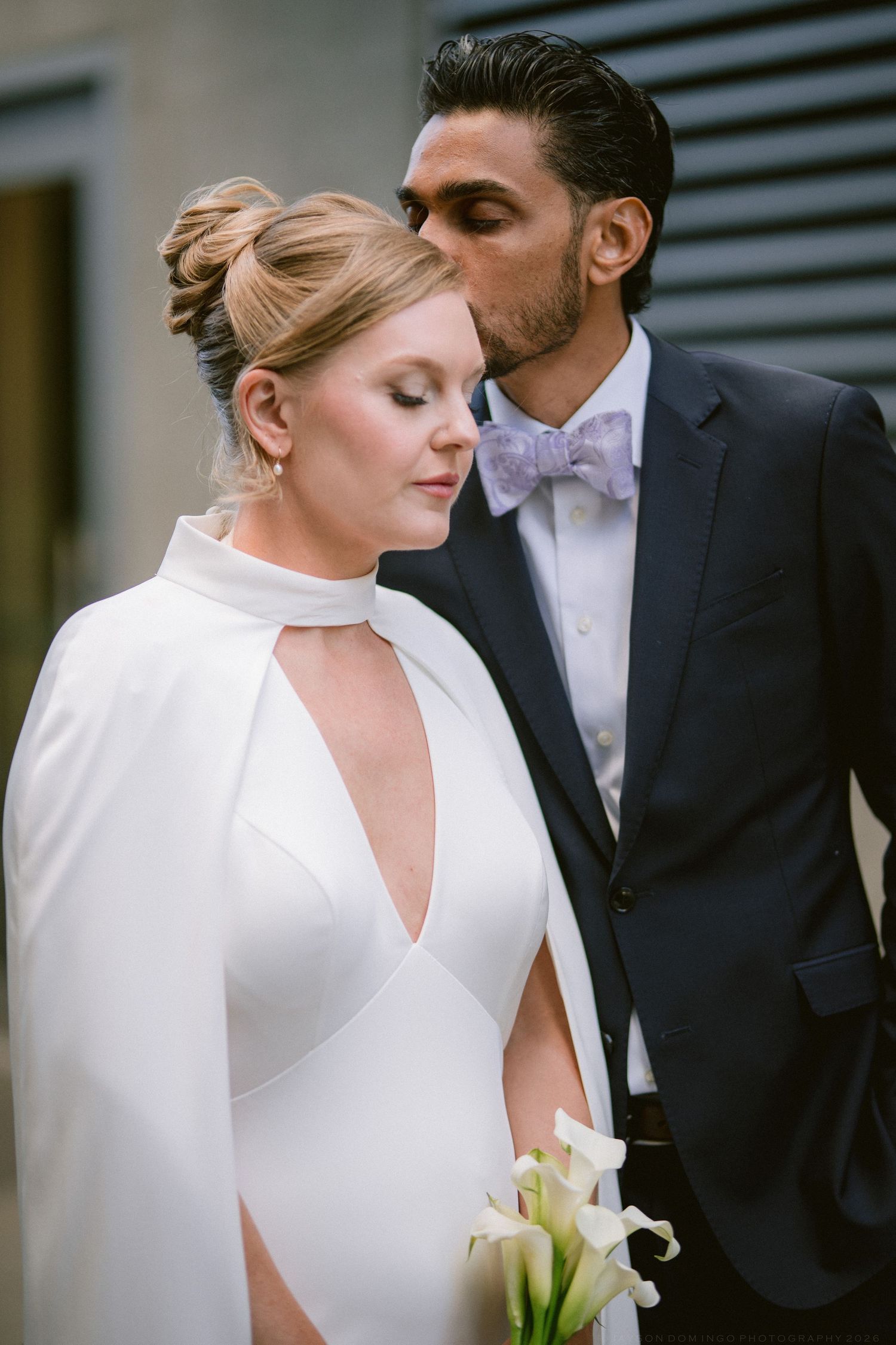 Groom in navy suit with lavender bow tie kissing bride wearing white cape wedding dress.