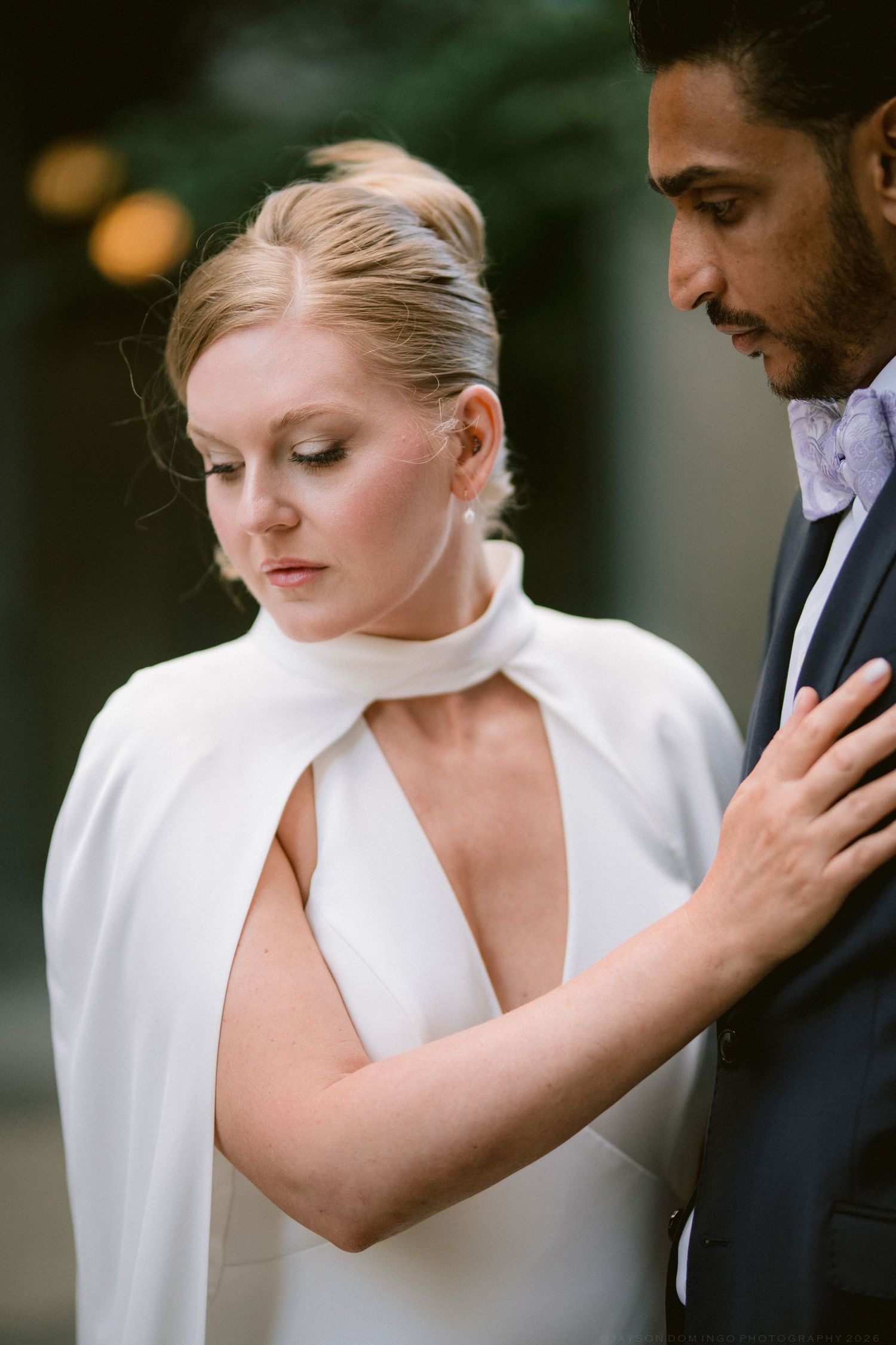 Bride in white cape wedding gown with groom in navy suit sharing an intimate moment outdoors.