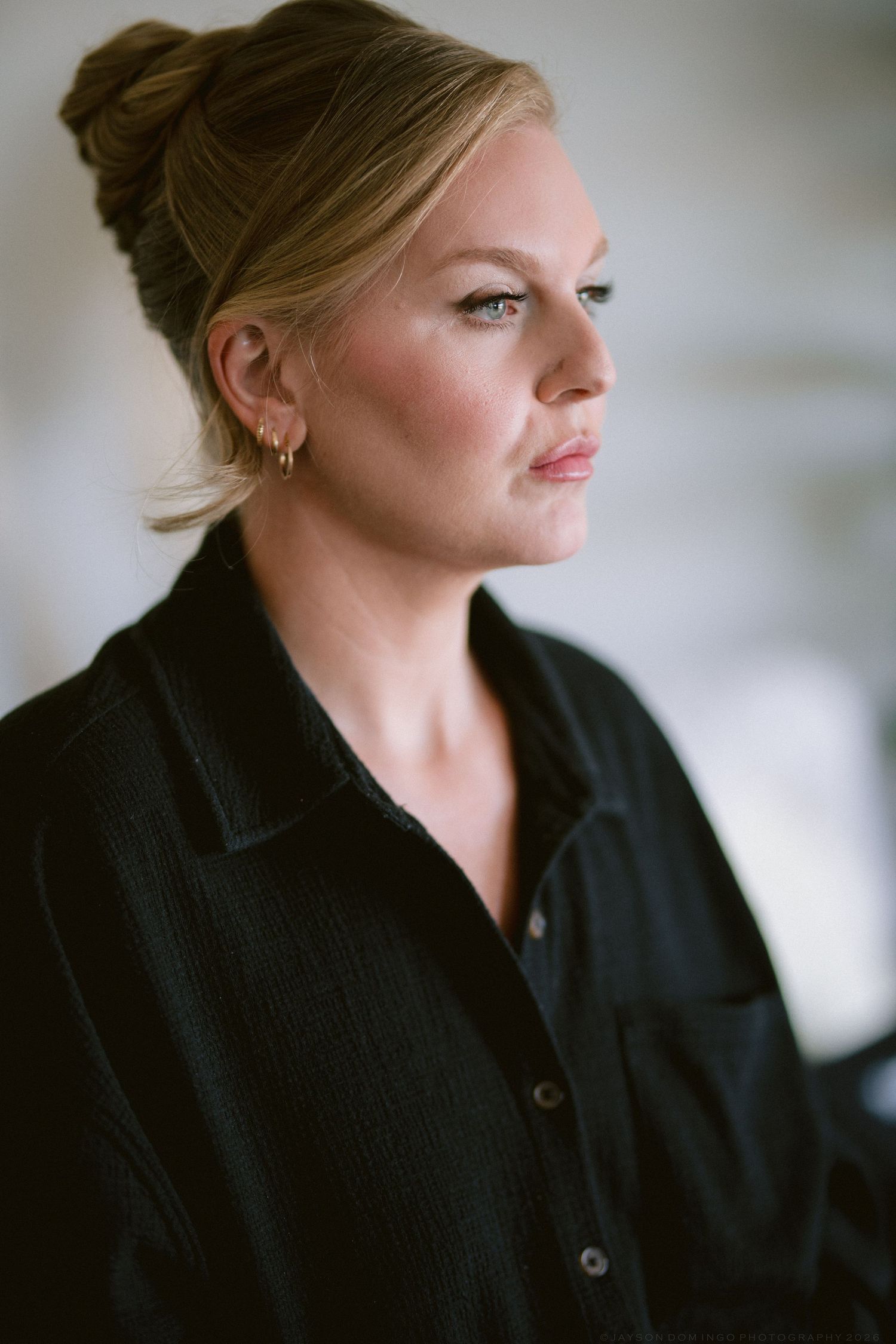 Blonde woman with updo hairstyle wearing black shirt gazes thoughtfully to the side in soft natural light.