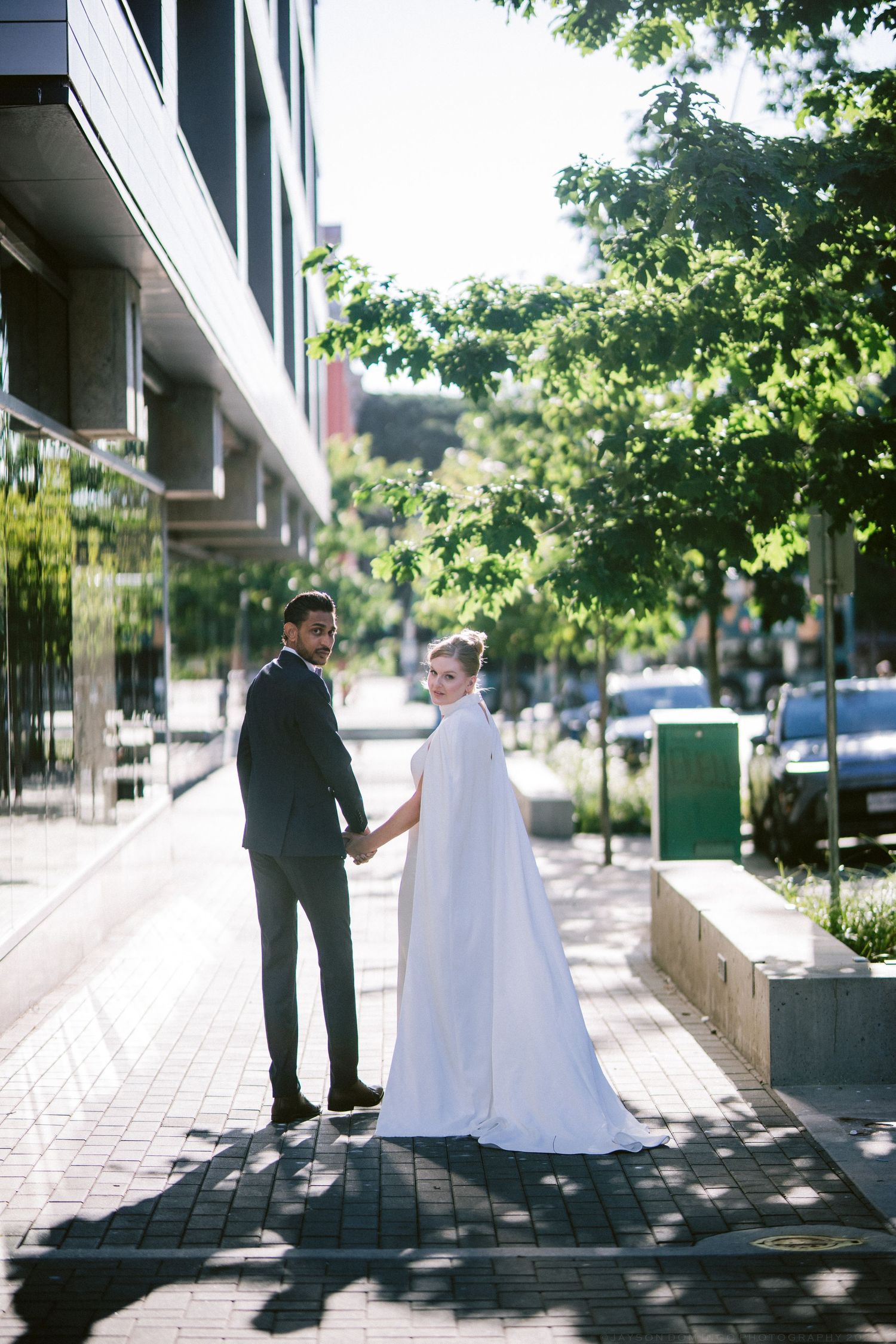 Newlywed couple holding hands in city street, bride wearing elegant white cape gown, groom in dark suit.