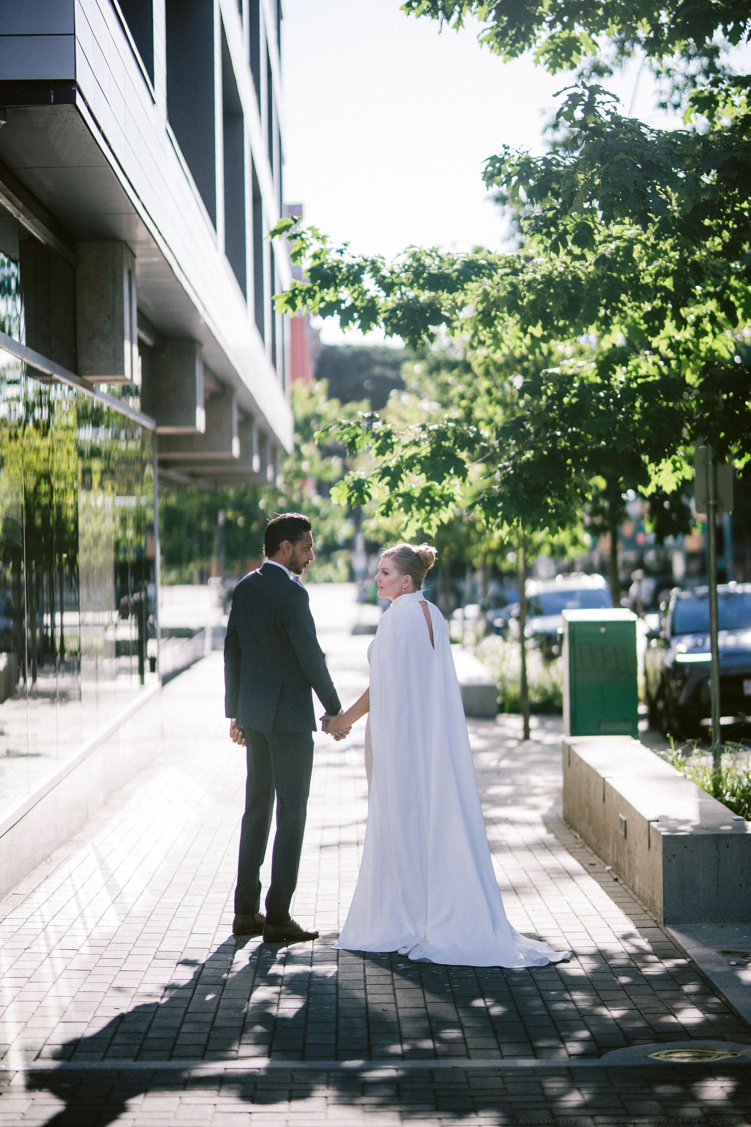 Bride and groom holding hands on urban sidewalk, bride wearing flowing white cape wedding gown.