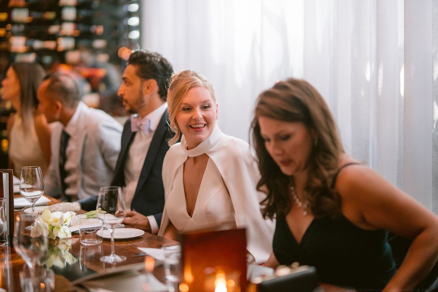 Smiling bride in white cape dress at wedding reception dinner table with guests.