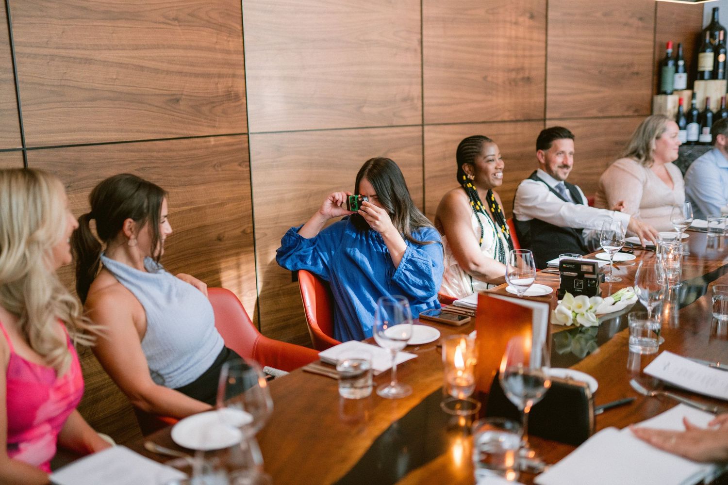 Guests laugh and enjoy wine at an elegant restaurant dinner party with warm wood-paneled walls.
