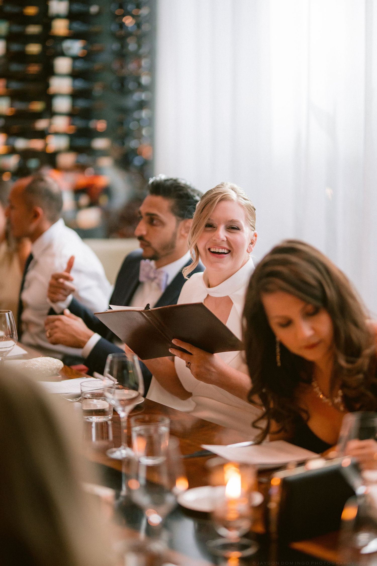 Smiling blonde bride in white dress reads menu at elegant restaurant wedding reception dinner.