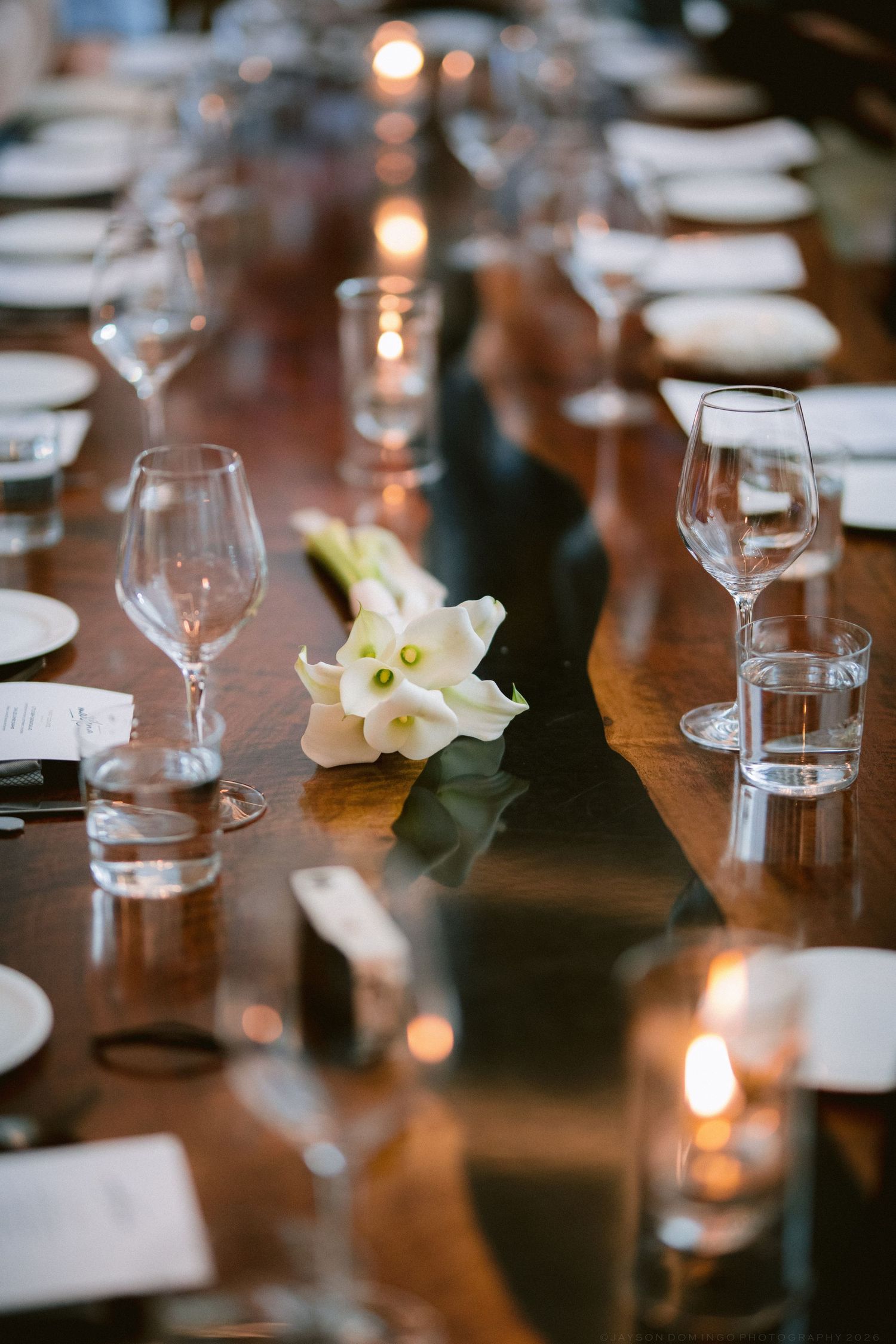 Elegant wedding reception table set with crystal glasses, white calla lilies, candles, and a dark runner on a wooden table.