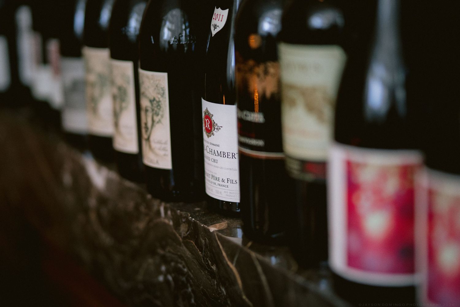 Close-up of wine bottles lined up on a stone shelf in a dimly lit cellar setting.