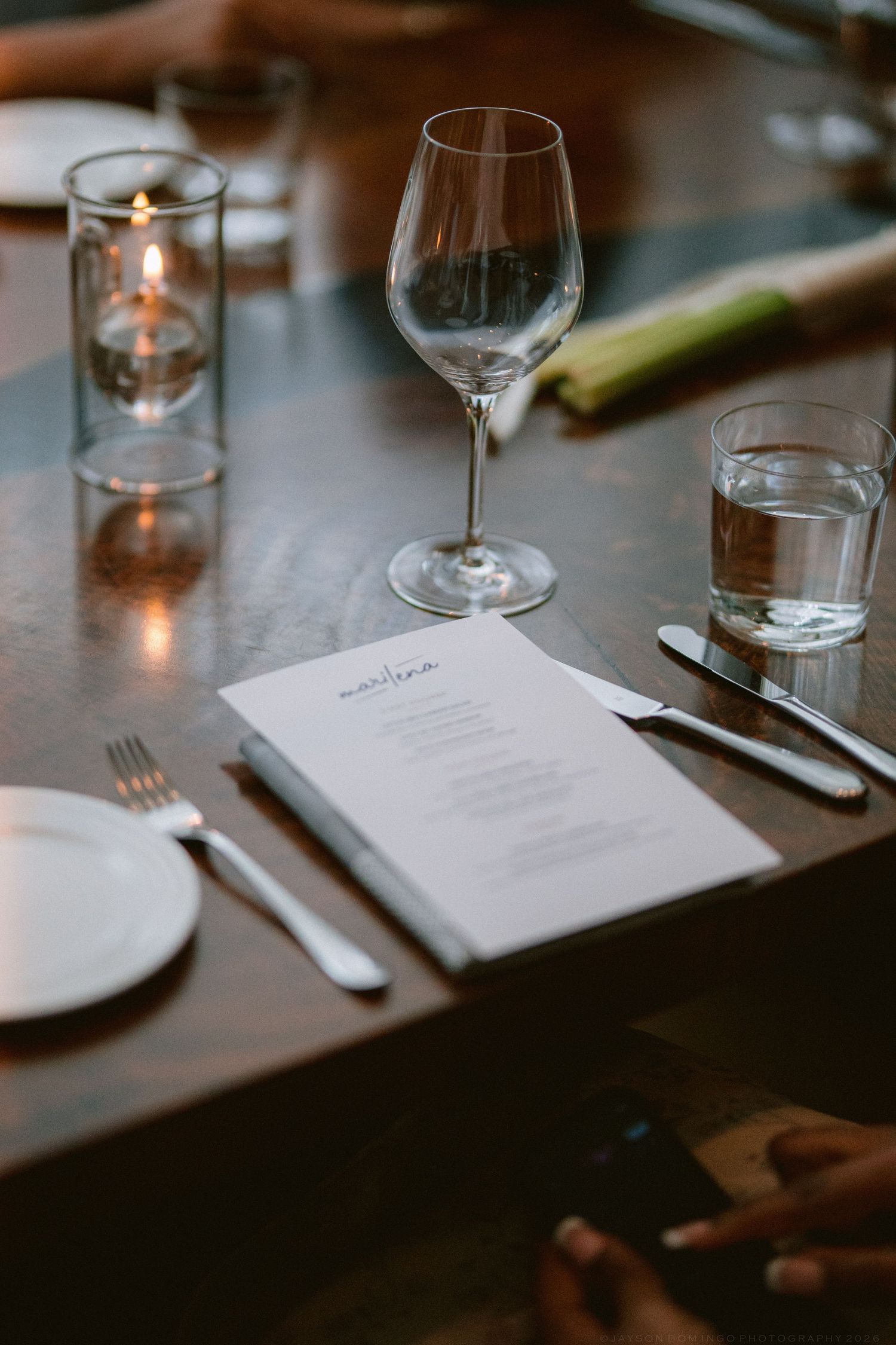 Elegant wedding reception place setting with wine glass, candles, menu card, and cutlery on dark table.